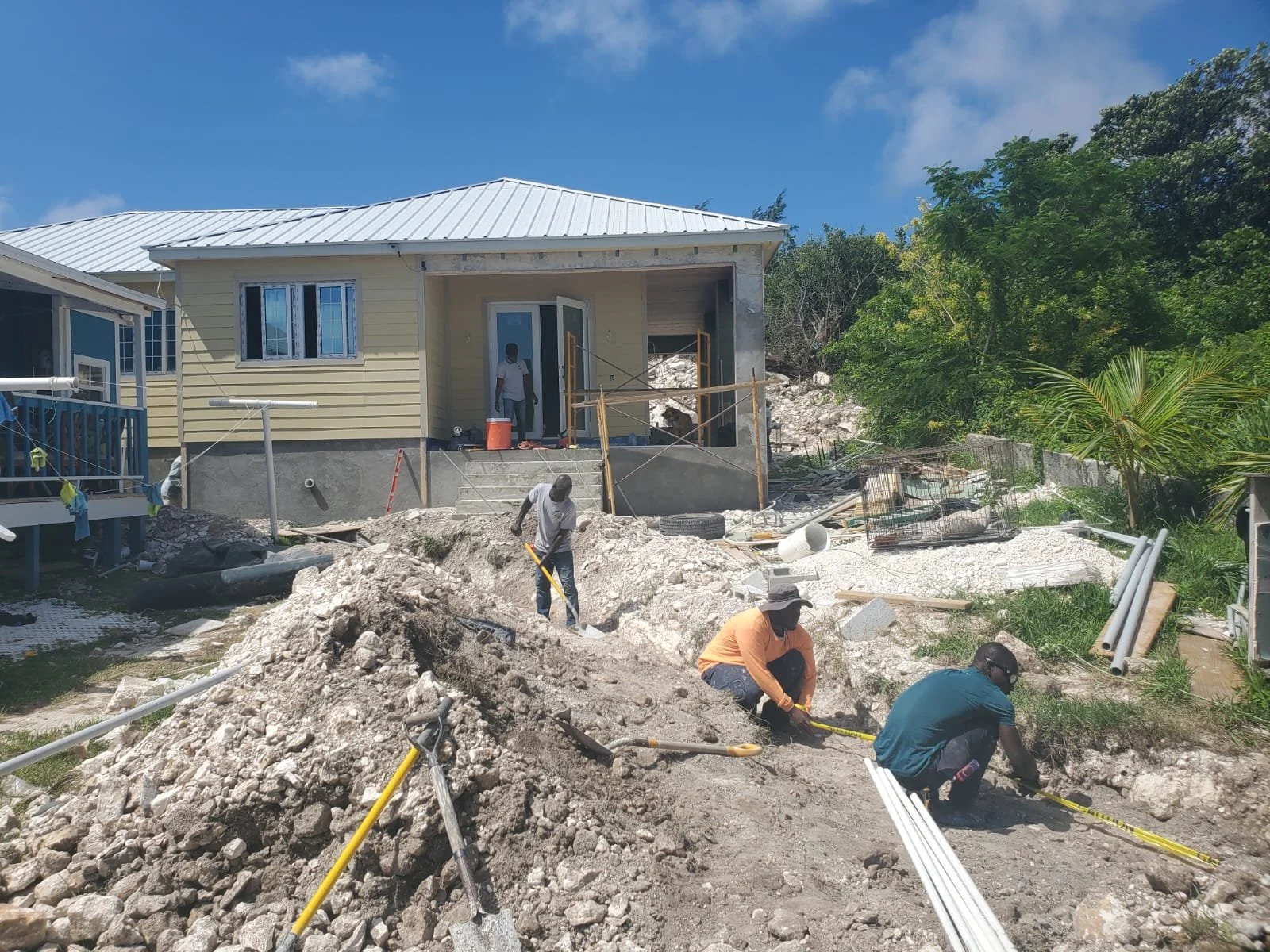 Construction workers leveling dirt in front of a house under renovation with a pile of dirt, tools, pipes, and vegetation around.