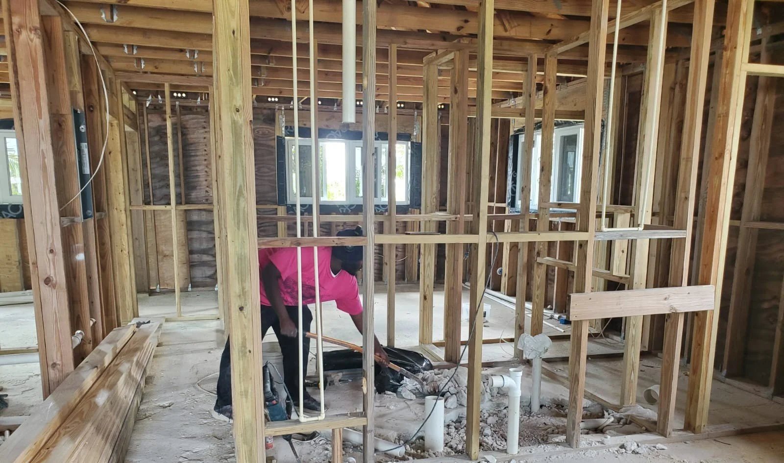 A man working on plumbing inside a house under construction, with exposed wooden framing and several windows.