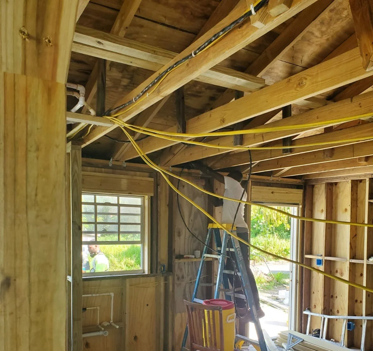 Interior view of a house under construction, showing exposed wooden framing, electrical wiring, a ladder, and construction worker."}