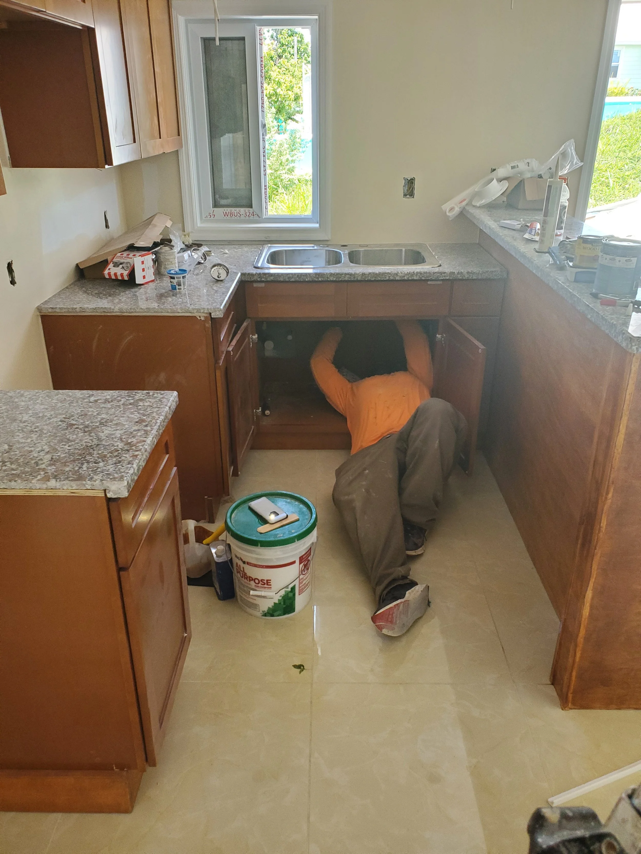 A person working underneath kitchen sink, partially under the countertop, with tools and supplies on the floor and countertops.