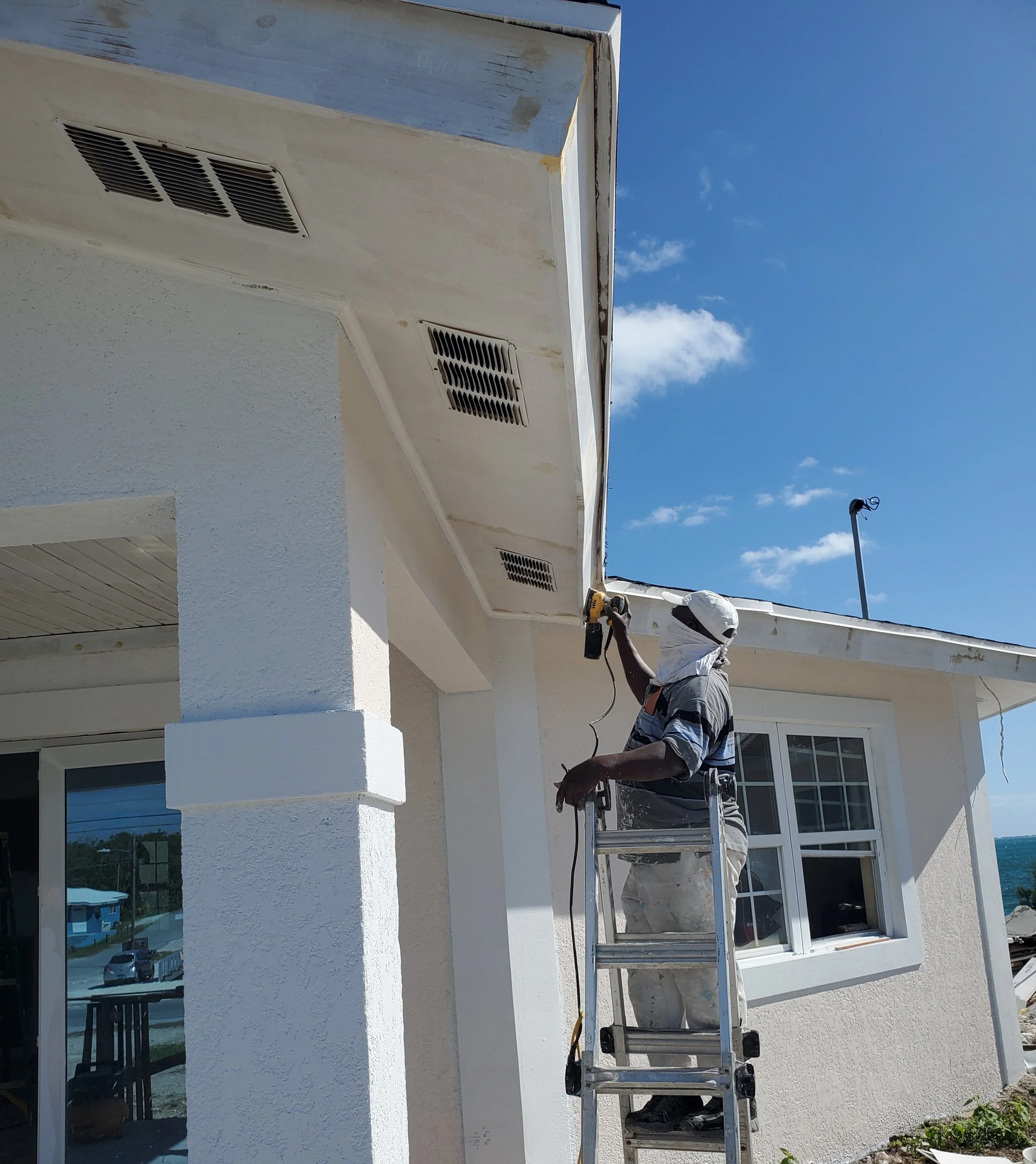 A worker on a ladder painting the exterior of a house under a blue sky with some clouds.