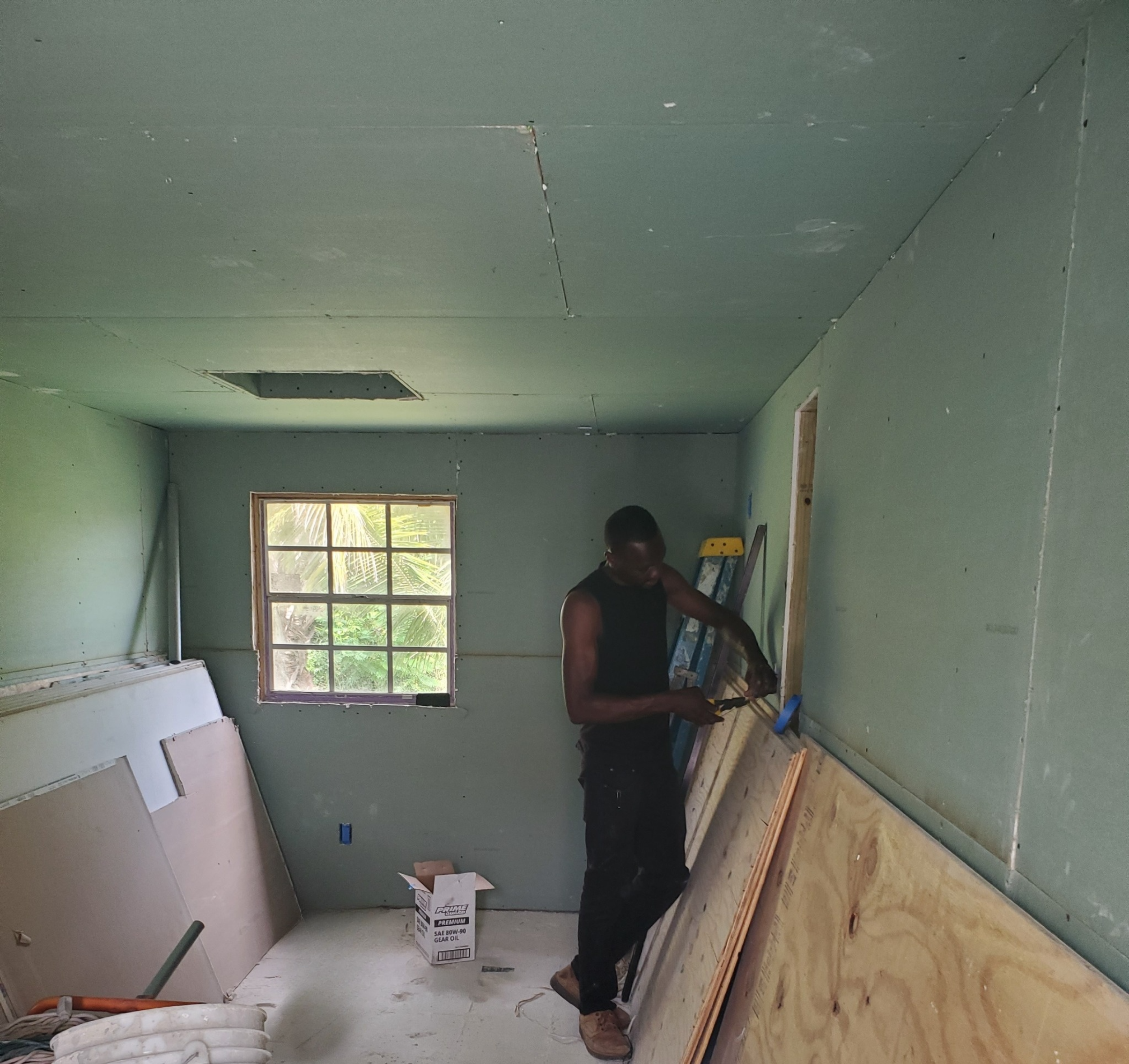 A man working on drywall installation in a partially finished room with green walls and a window, construction tools and materials around.