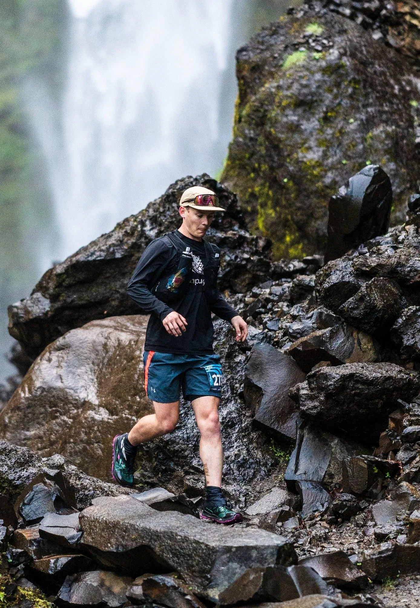 A male trail runner wearing a beige cap, black long-sleeve shirt, blue shorts with orange details, and trail shoes, running on a rocky, wet trail near a waterfall. Gorge Waterfalls