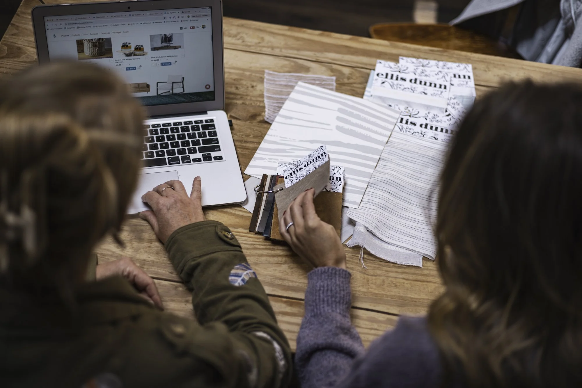 Two women sitting at a wooden table, looking at fabric samples and browsing an online furniture website on a silver laptop.