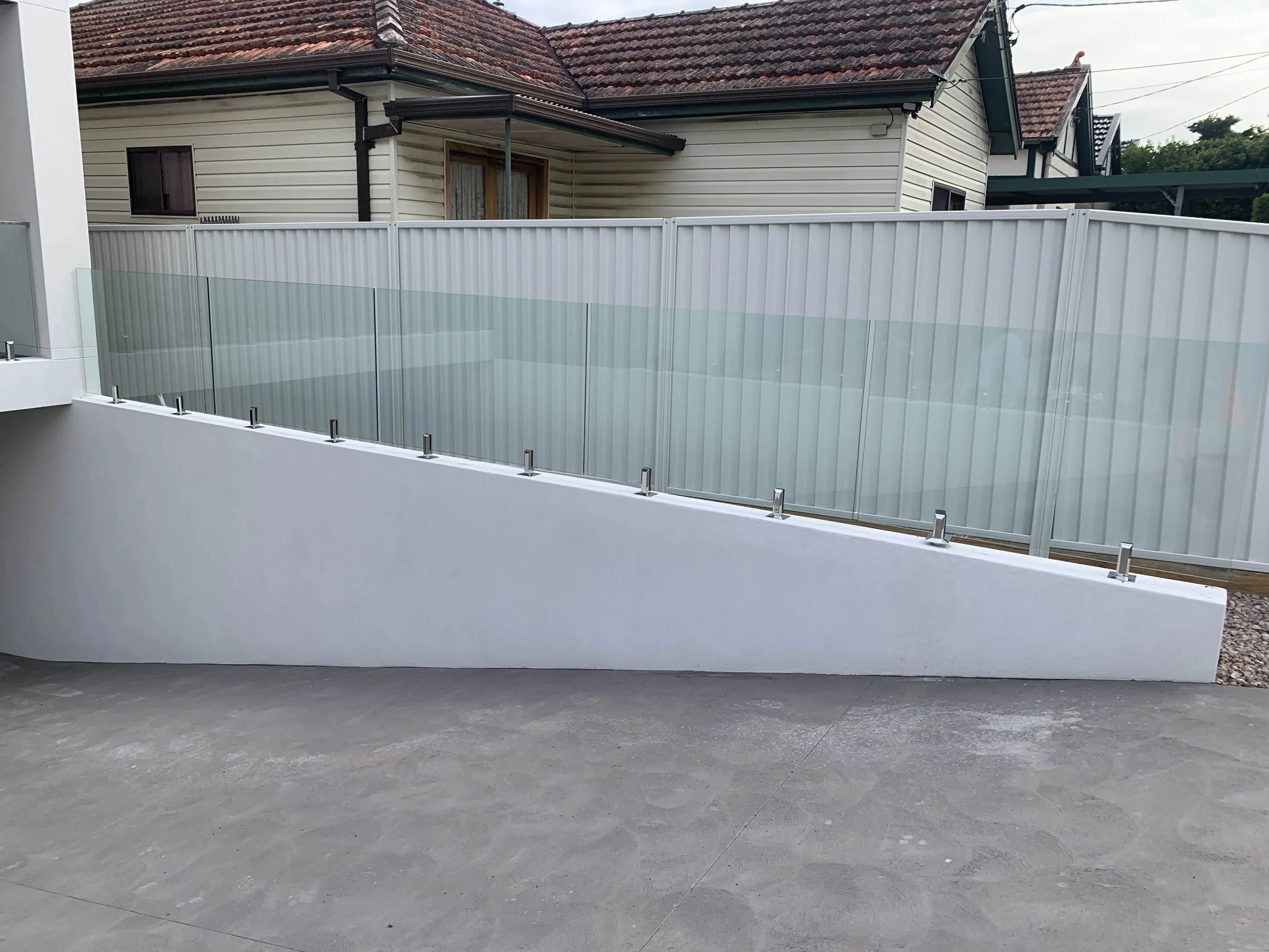 Glass fence on a sloped concrete surface in front of a house with gray siding and red-tiled roof.