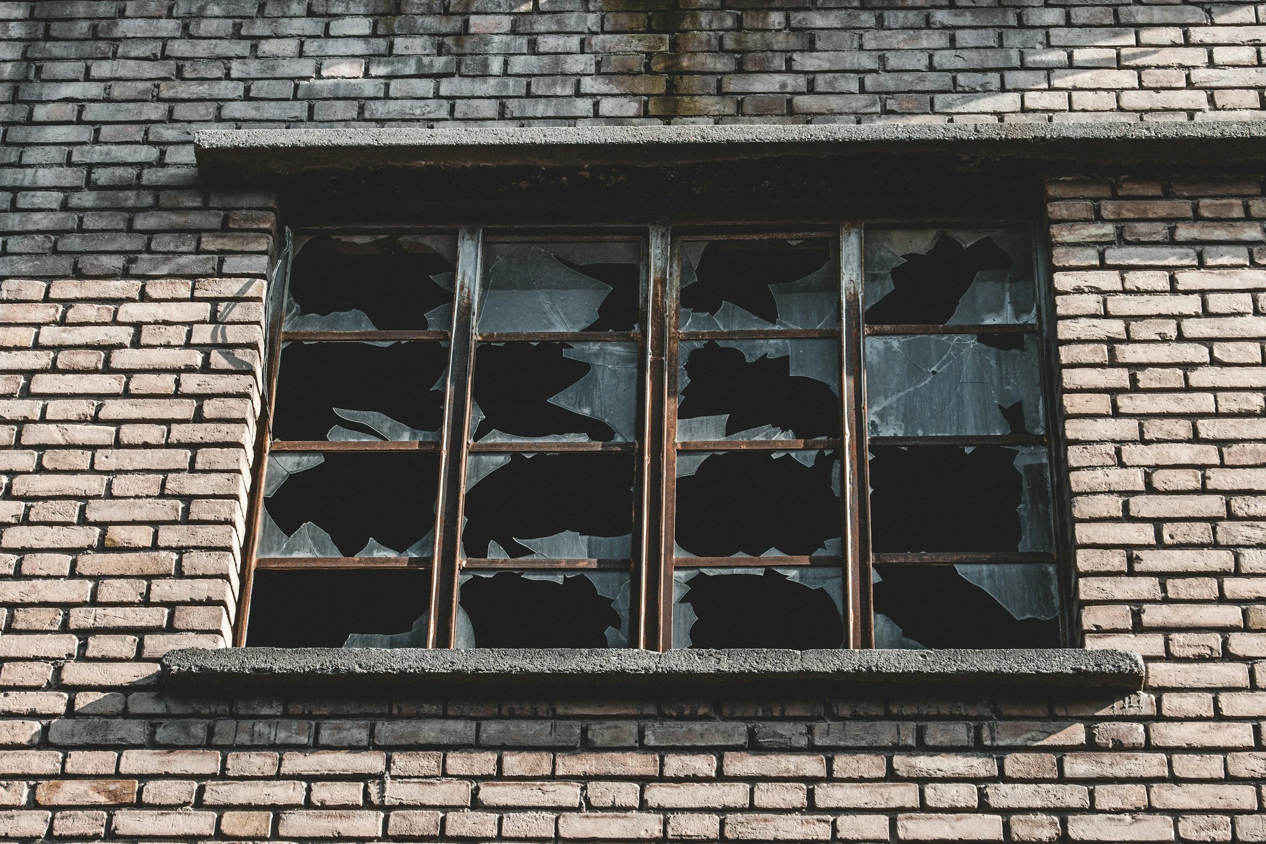 A black metal window frame with broken and missing glass, some pieces still attached, and spider webs on the edges.