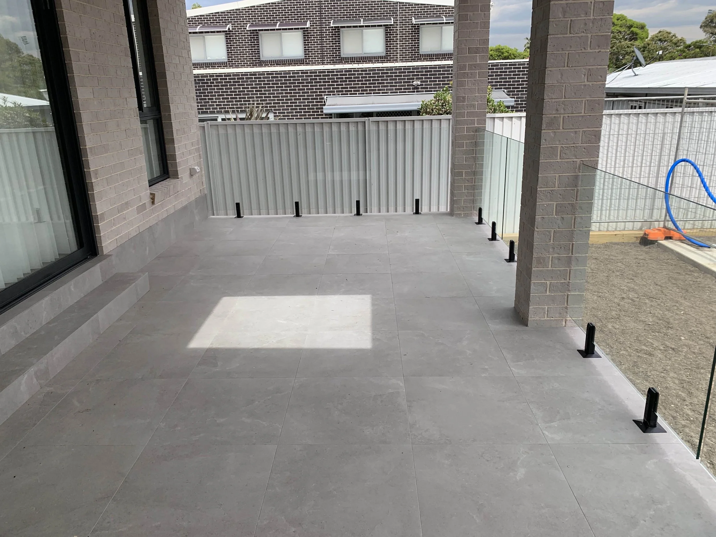 Modern outdoor patio with gray floor tiles, brick pillars, glass railing, and view of adjacent buildings.