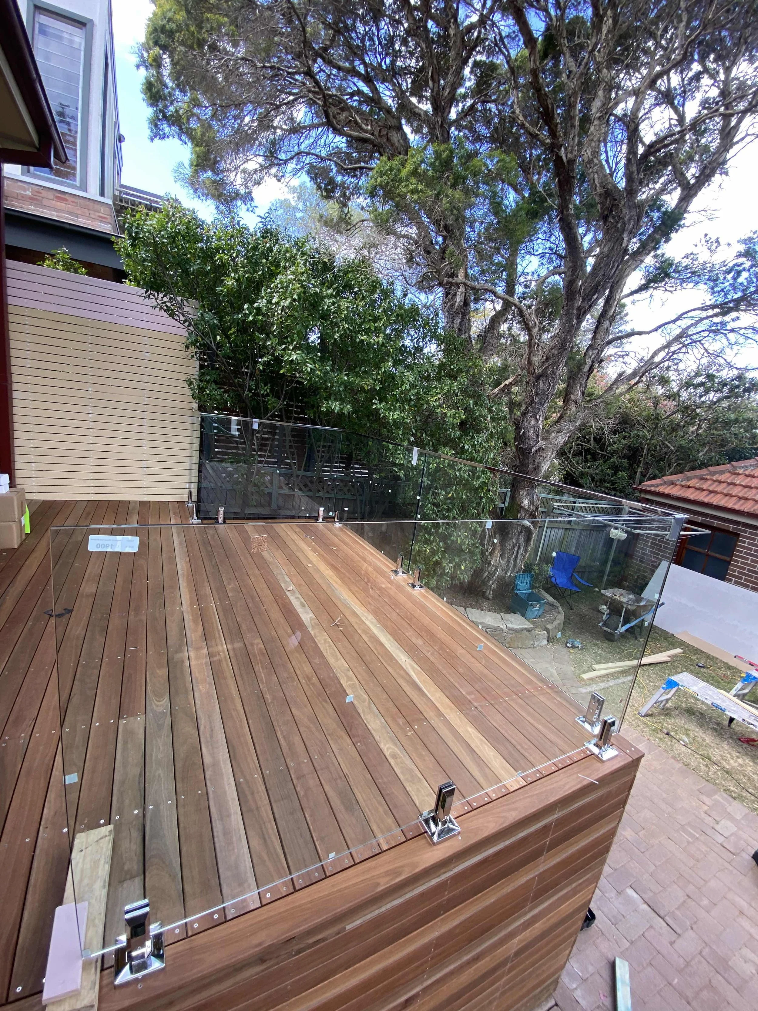 A wooden deck with a glass railing, surrounded by trees and outdoor items like chairs and a wheelbarrow. The setting appears to be a backyard of a house.