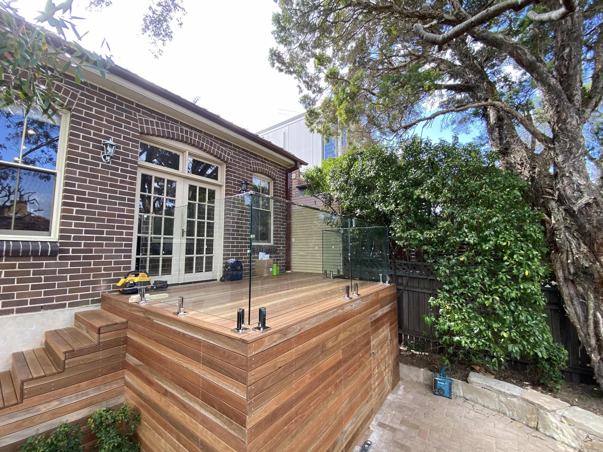 A wooden patio with glass railing attached to a brick house, surrounded by trees and foliage.
