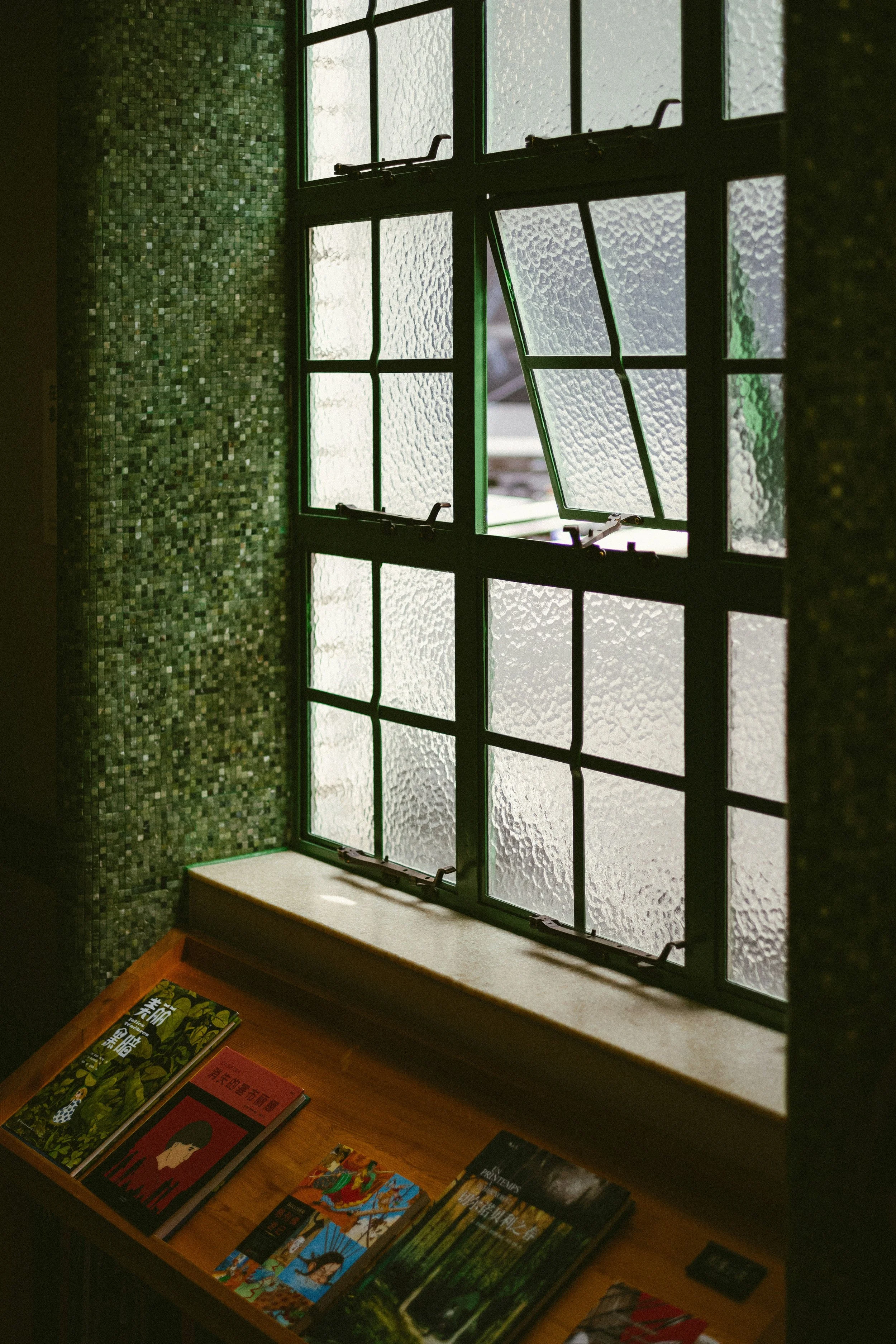 Open textured glass window with a wooden ledge holding books inside a room with green mosaic tiled wall.