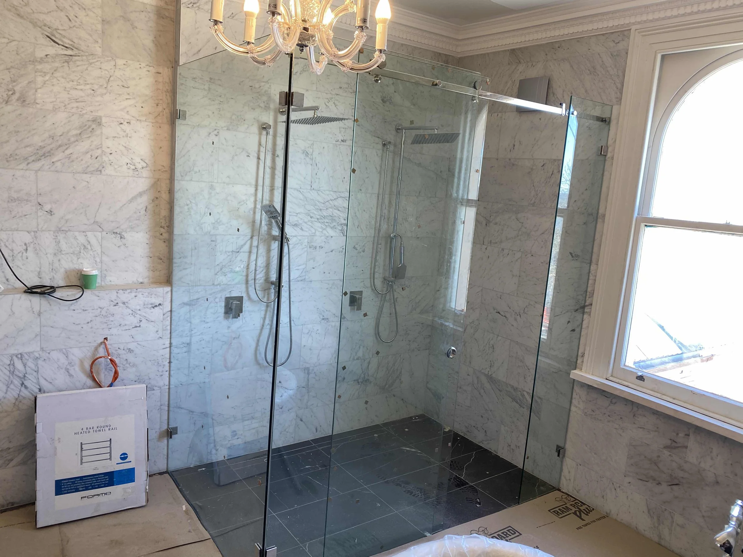 Bathroom under renovation with a glass-enclosed shower, marble walls, and a chandelier above. A window is on the right side, allowing natural light to enter the room.