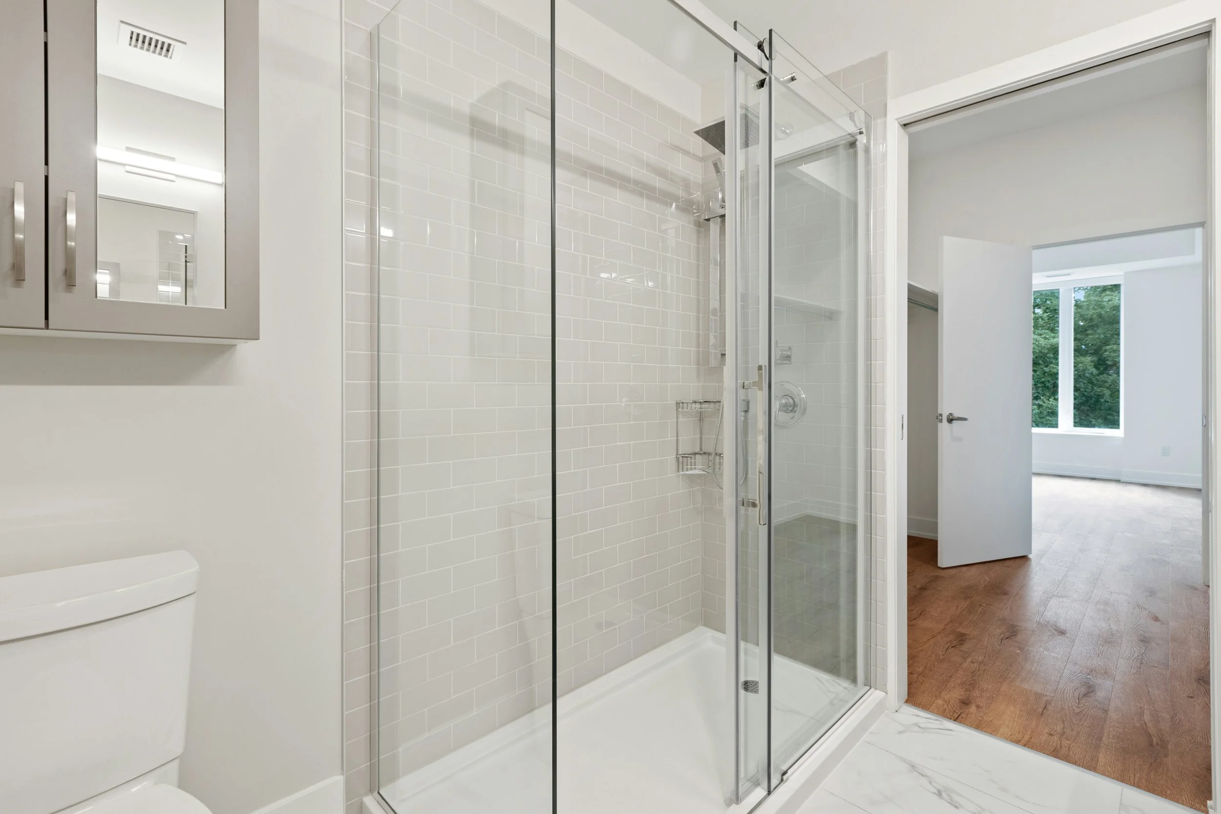 Modern bathroom with a glass-enclosed shower, white tile walls, a toilet, and a gray wall-mounted cabinet. An open door reveals a bright room with large windows and wooden flooring.