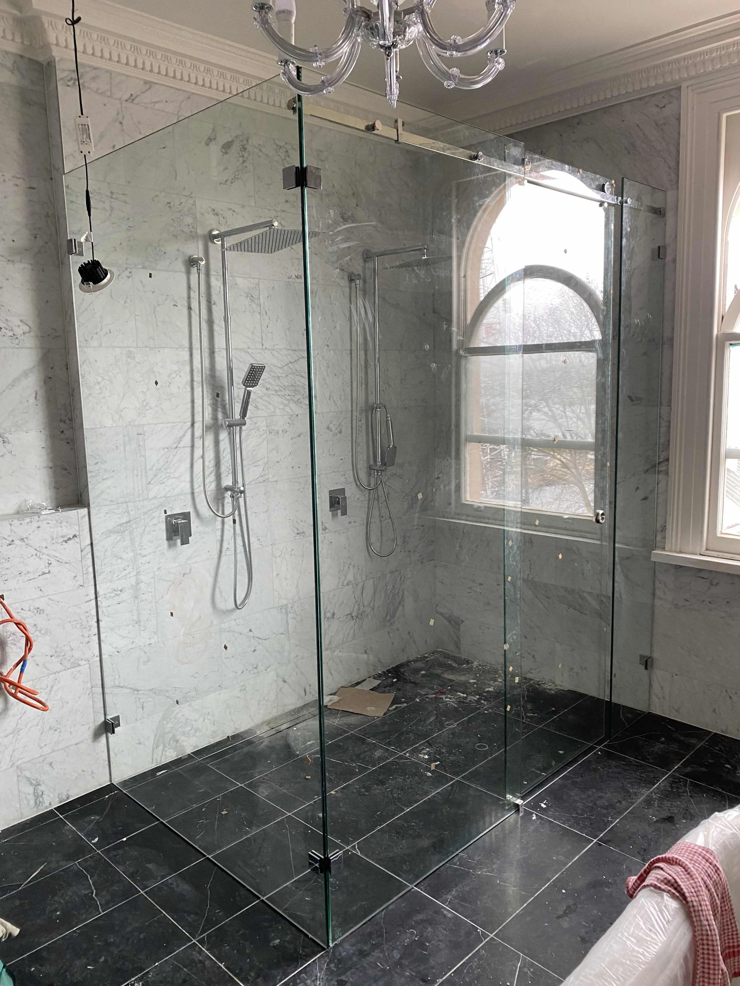 Luxury bathroom with a glass shower enclosure, marble walls, and black tiled floor; features a rainfall showerhead and handheld shower, with a chandelier overhead.