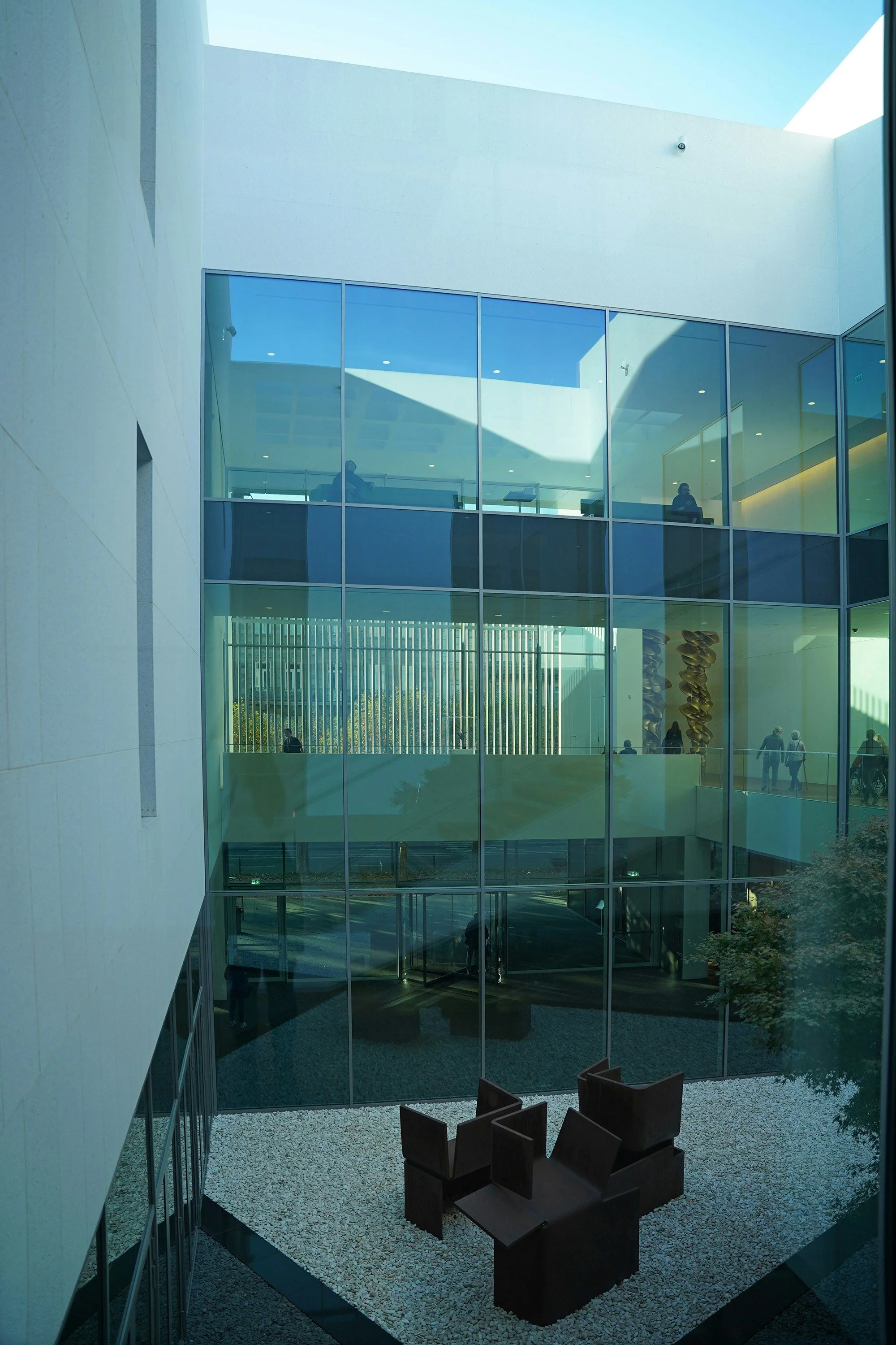 Interior view of a modern office building with a central courtyard, glass walls, and a seating area of four dark chairs on white gravel, with people visible inside and outside the building.