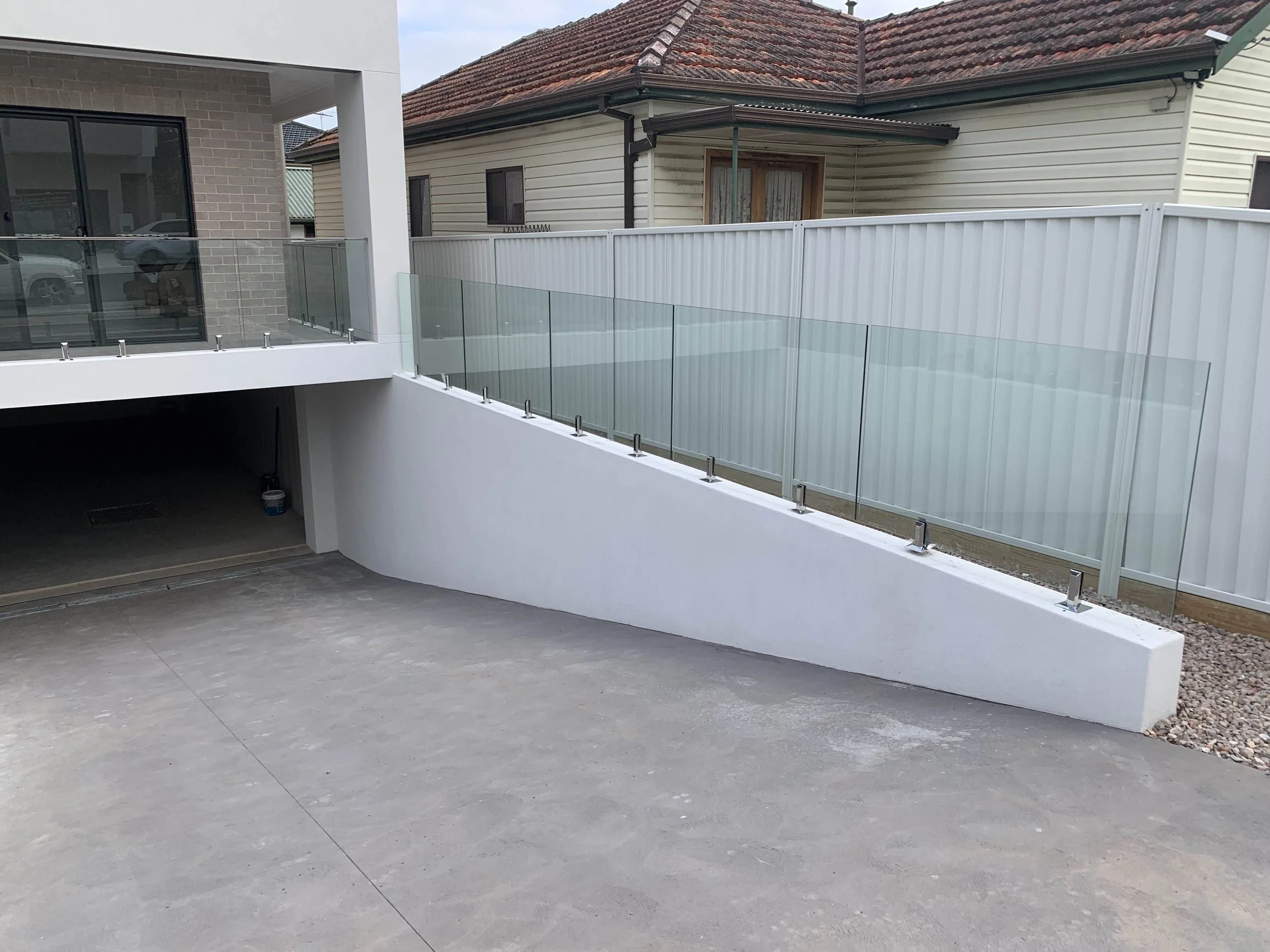 Residential driveway with glass railing and white retaining wall, leading to a garage entrance, with neighboring houses visible in the background.