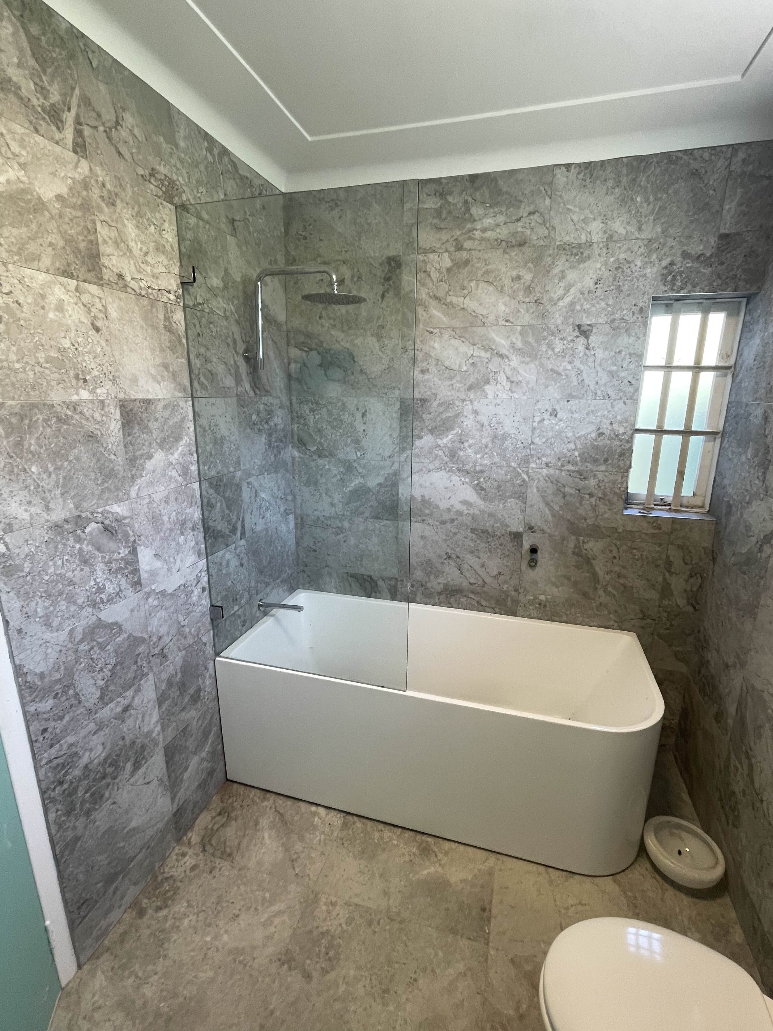Modern bathroom with gray marble tiles, a white bathtub, a glass shower screen, a small window with bars, and a white toilet.