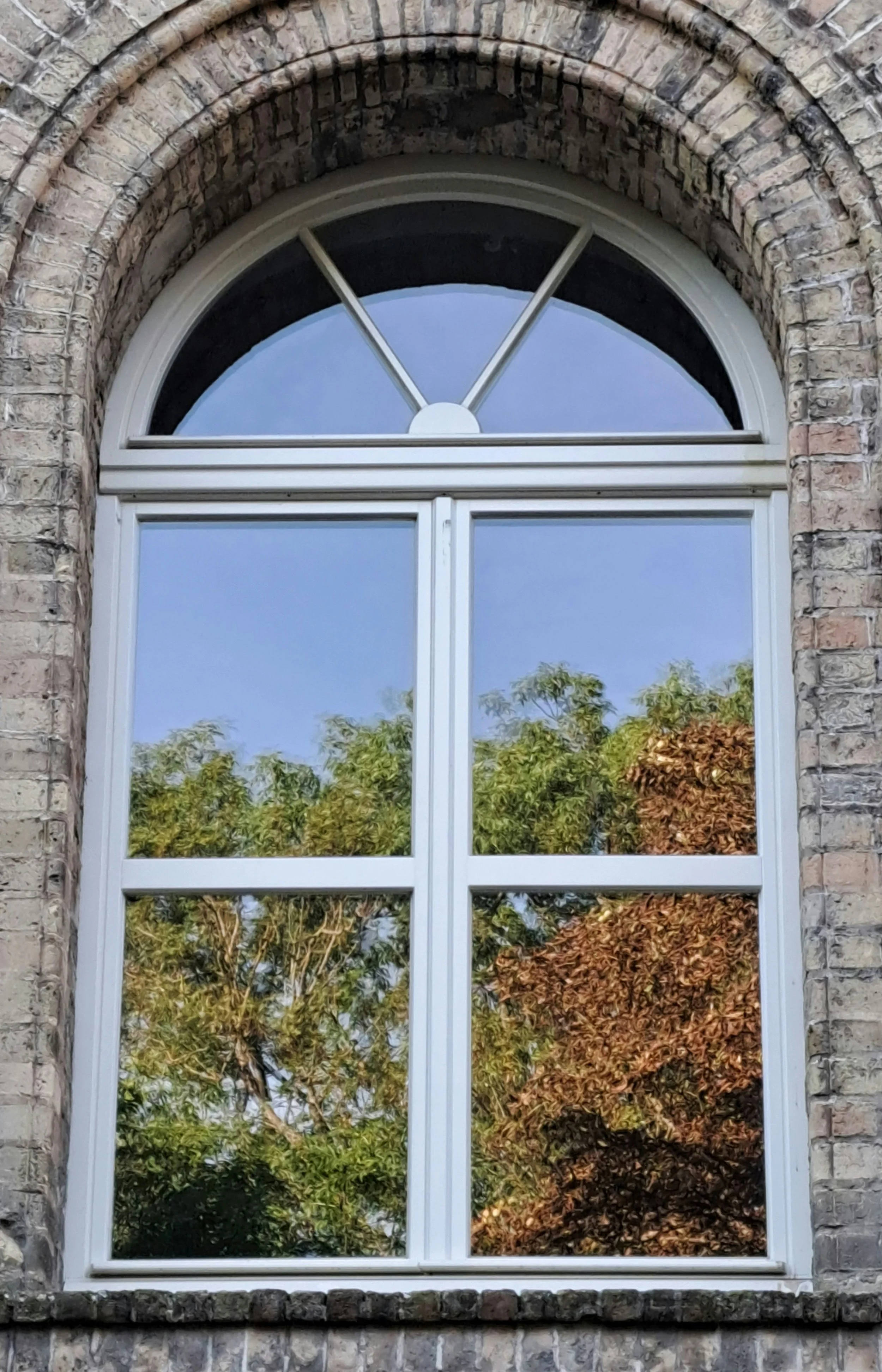 A brick building window with a semi-circular top mirror reflecting blue sky and green and brown autumn trees.