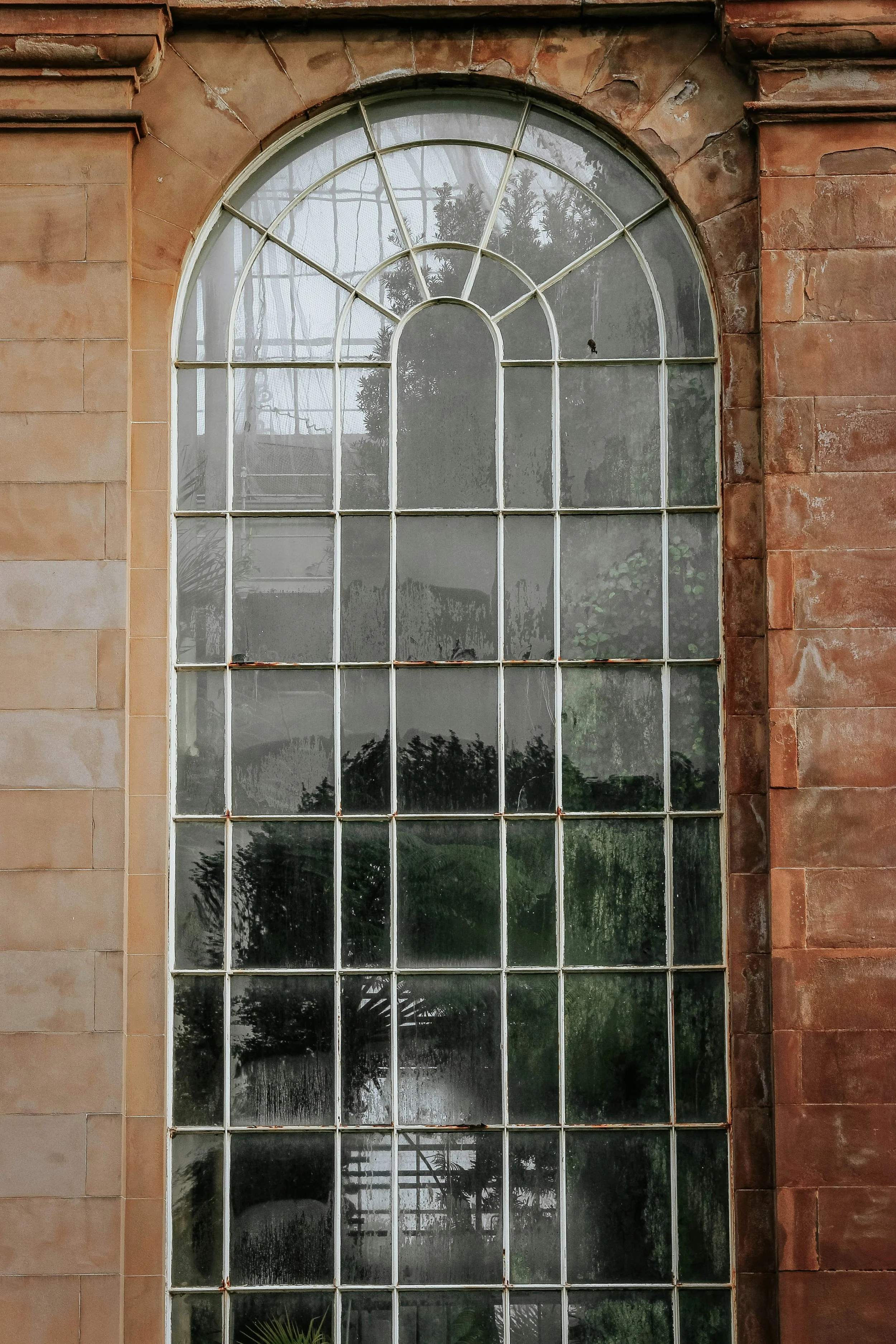 Large arched window with multiple panes, set in a brick and stone wall, with reflections of trees and sky visible on the glass.