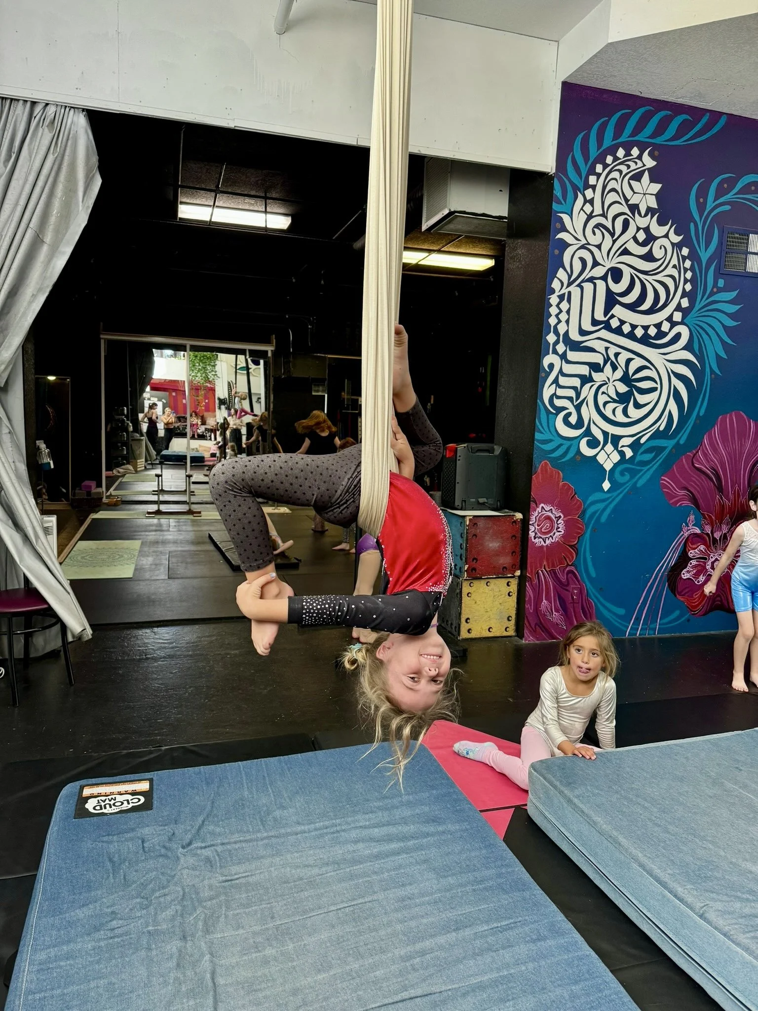 A young girl practicing aerial silk yoga upside down, smiling, in a gym, with other children in the background.