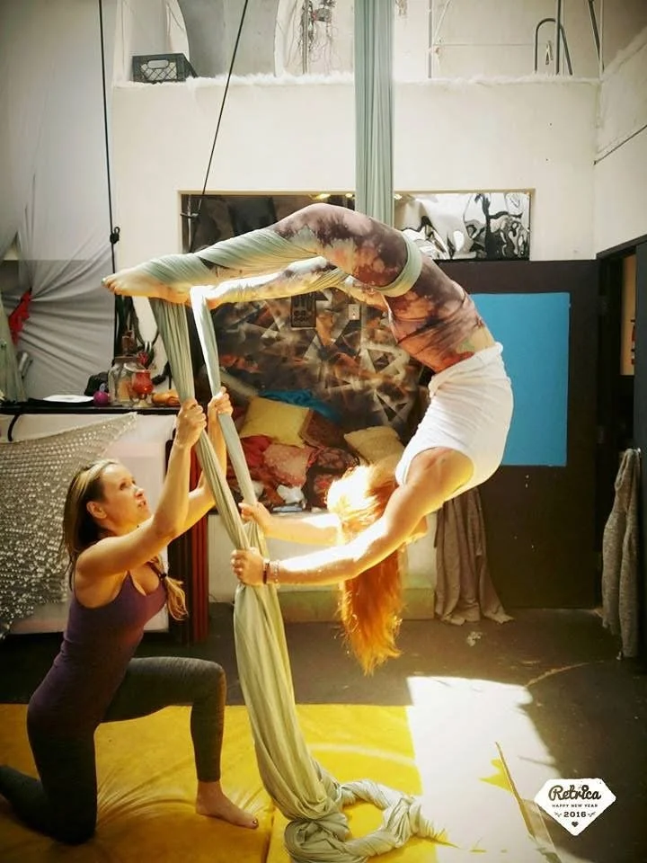 Two women practicing aerial silk acrobatics indoors, one woman assisting the other with an inverted pose.