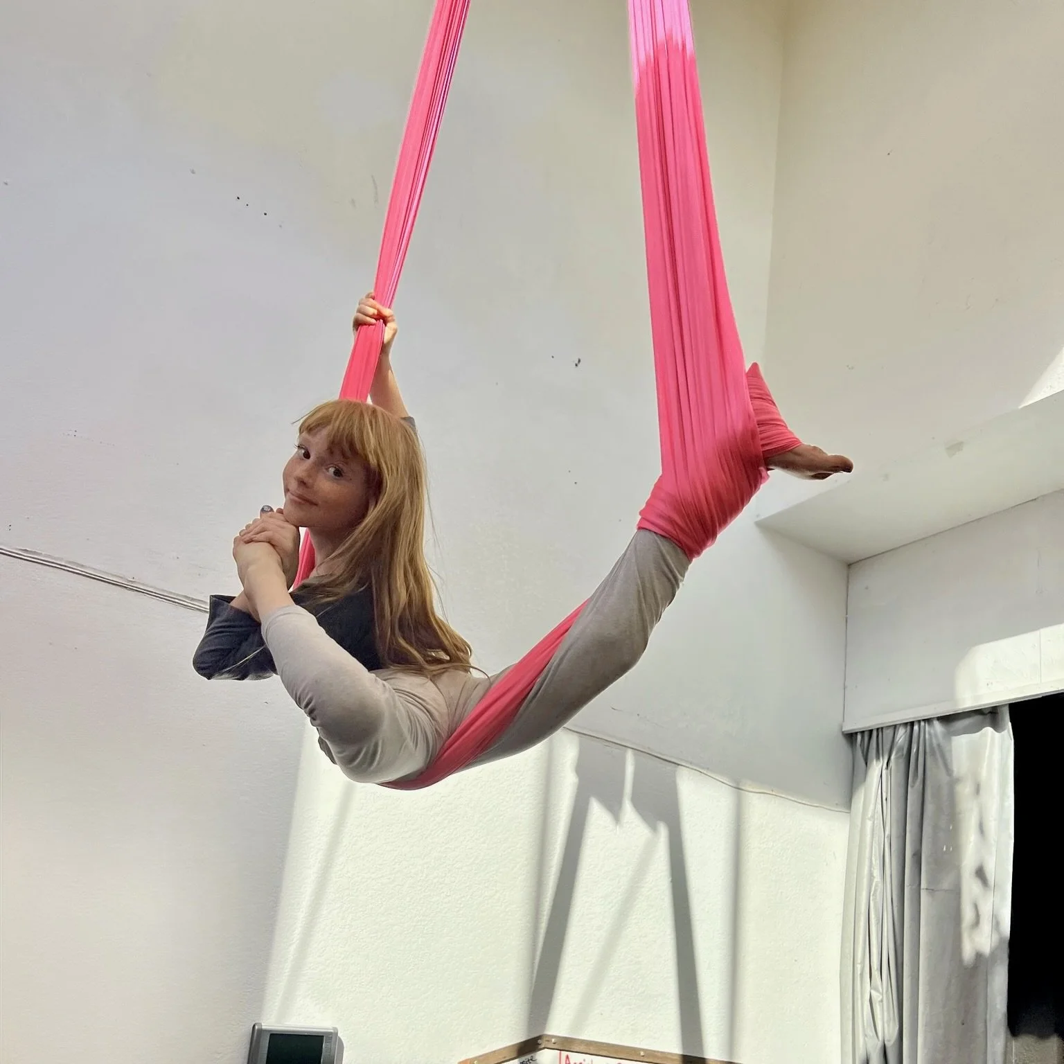 A woman with red hair lies in an aerial silk hammock hanging from the ceiling, holding onto the silk with one hand, while her legs are wrapped in pink fabric. She rests her chin on her hands and looks at the camera.