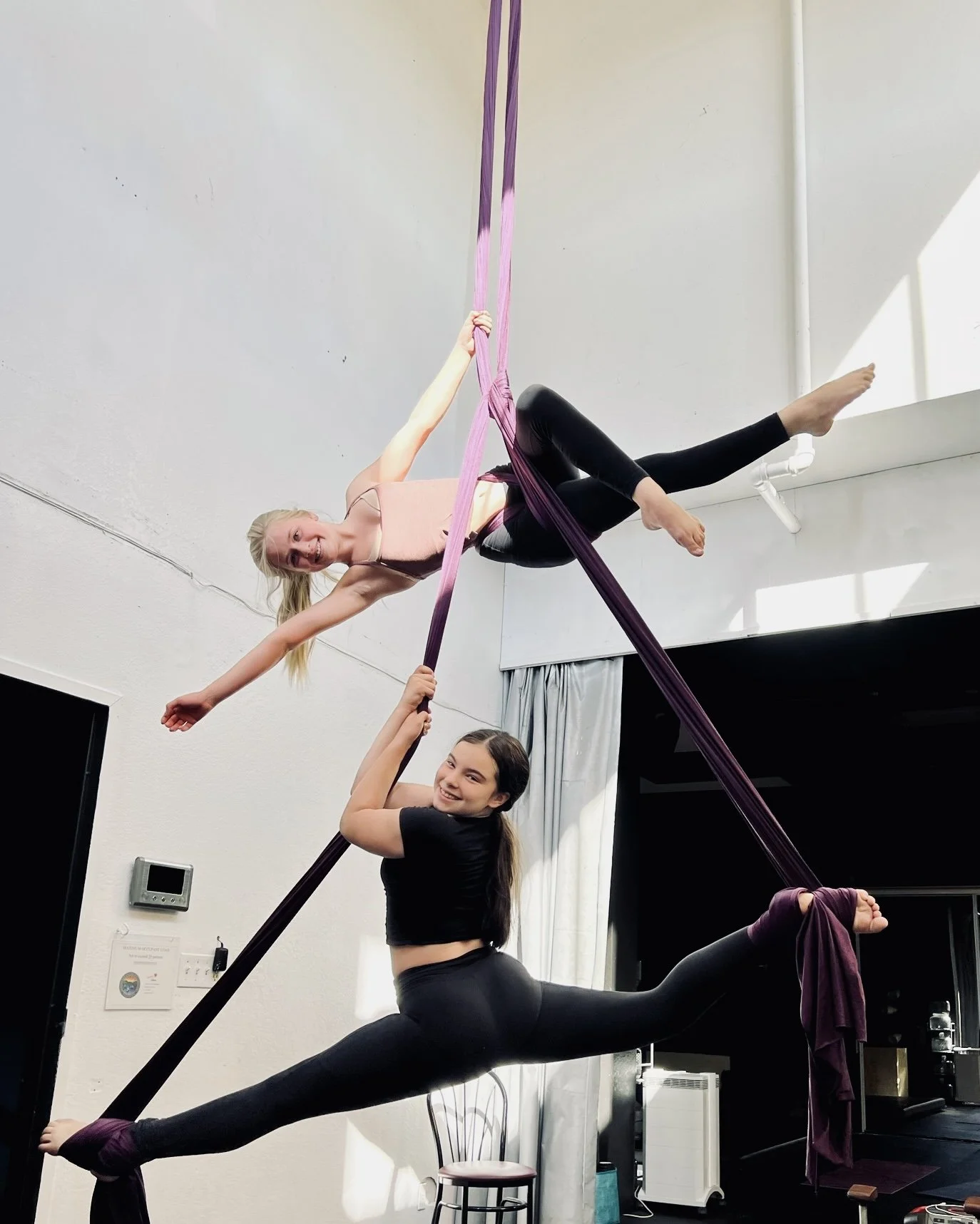 Two women practicing aerial silk performance indoors, one hanging from pink silk and the other on the floor holding the silk, both smiling.