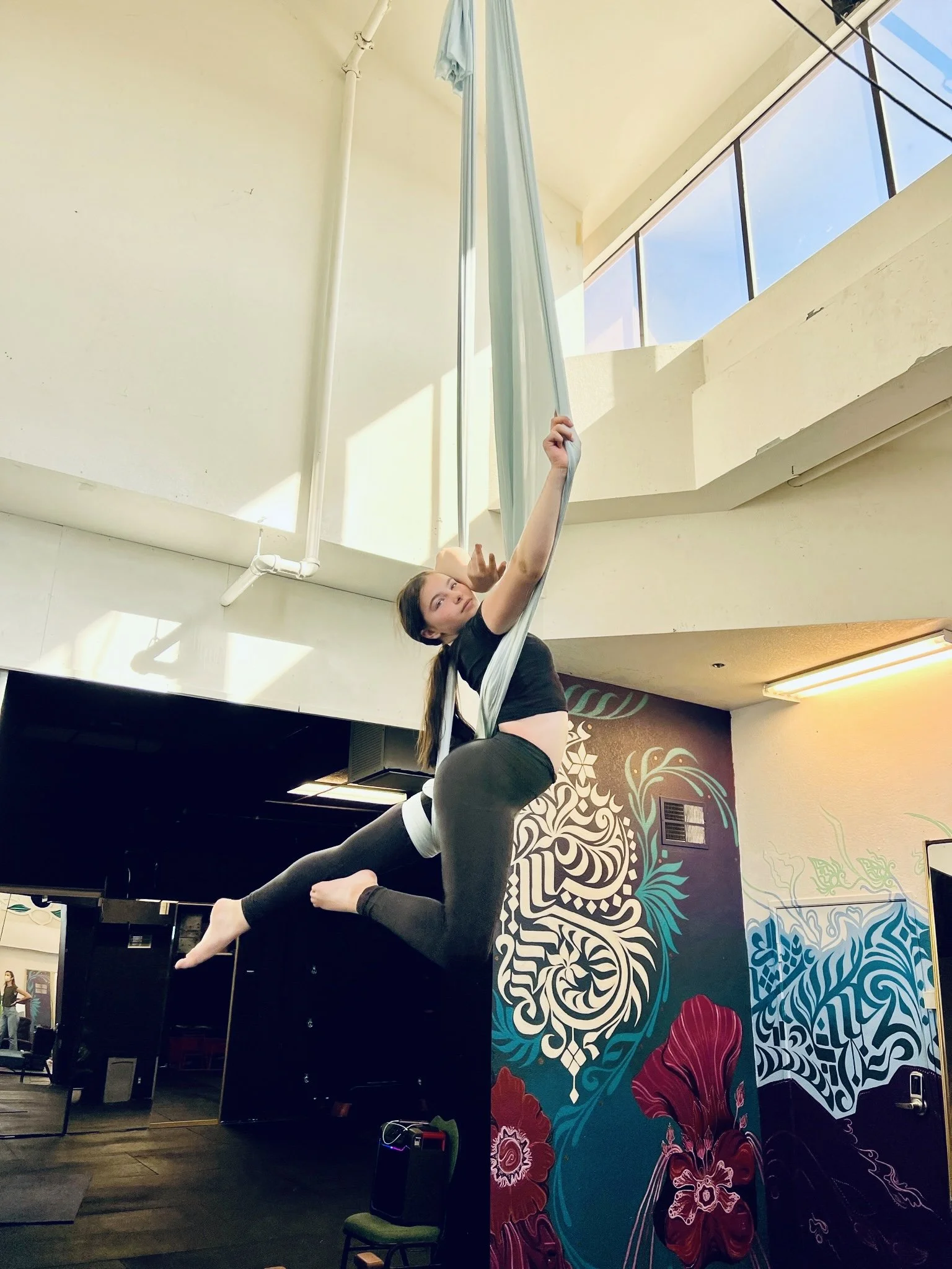 Woman practicing aerial silk acrobatics indoors, hanging from blue fabric, with a colorful mural on the wall behind her.
