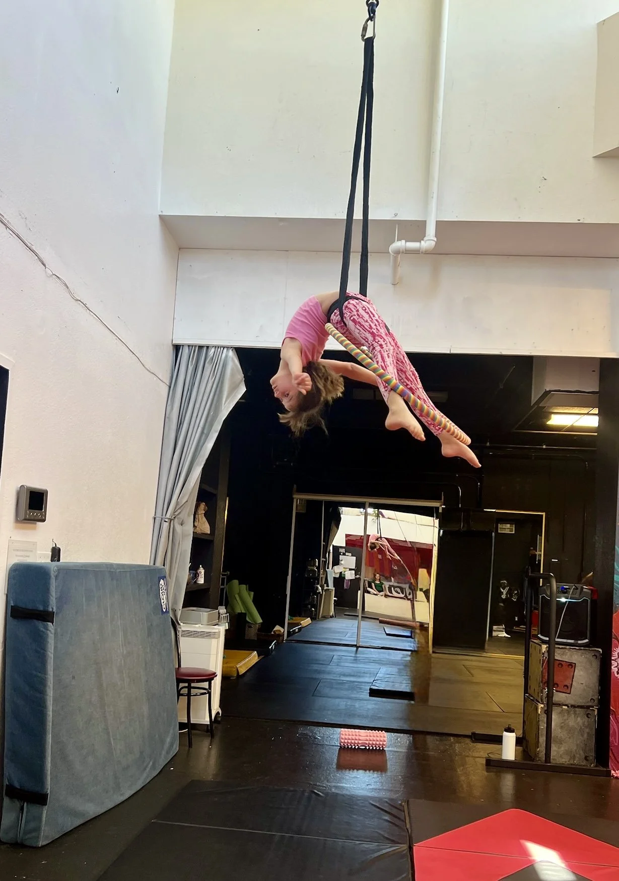 A young girl hanging upside down on a gymnastics ribbon at an indoor gymnasium, with gym equipment and mats around.