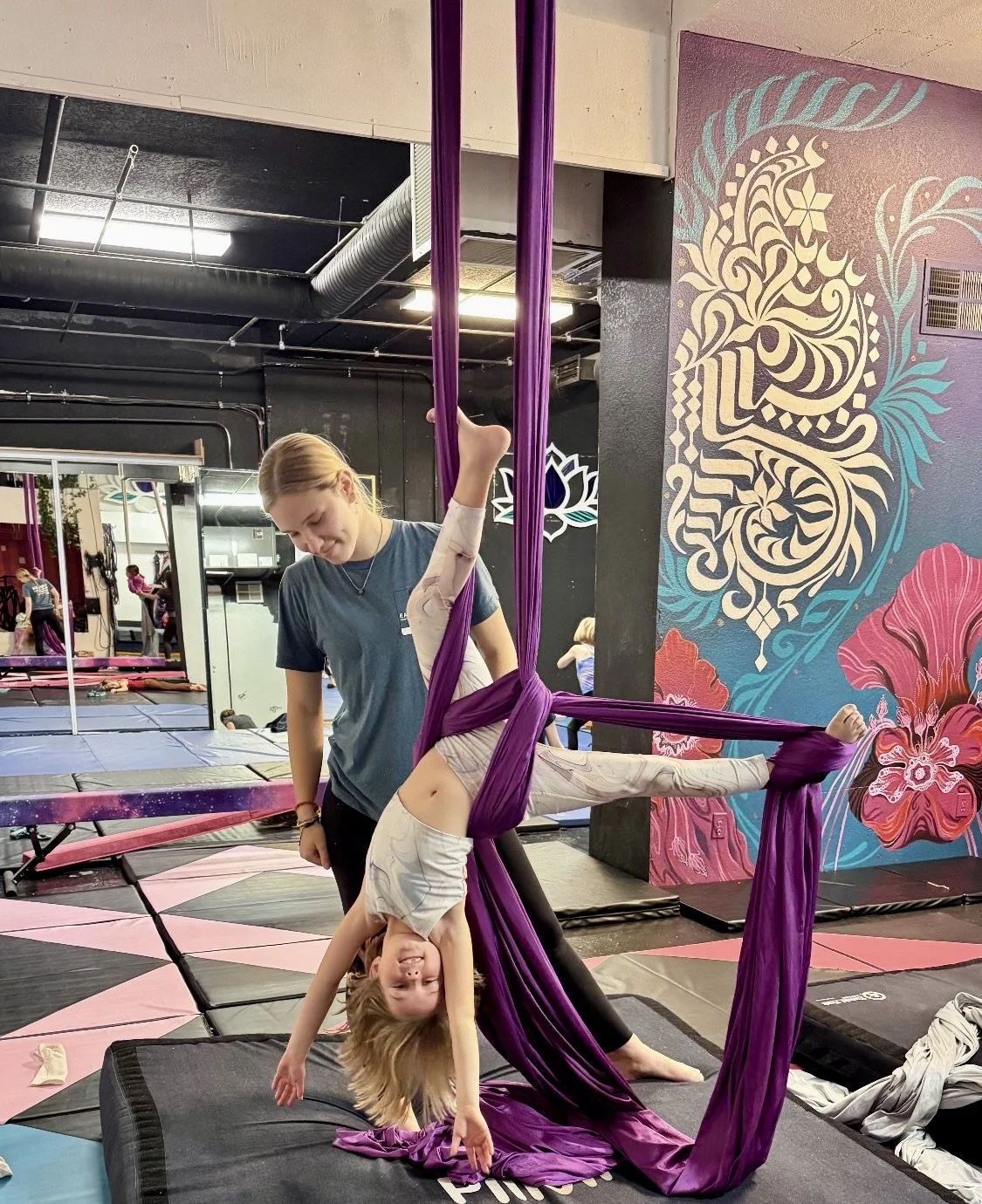 A young girl practicing aerial silk gymnastics with a trainer in a dance studio with colorful wall art.