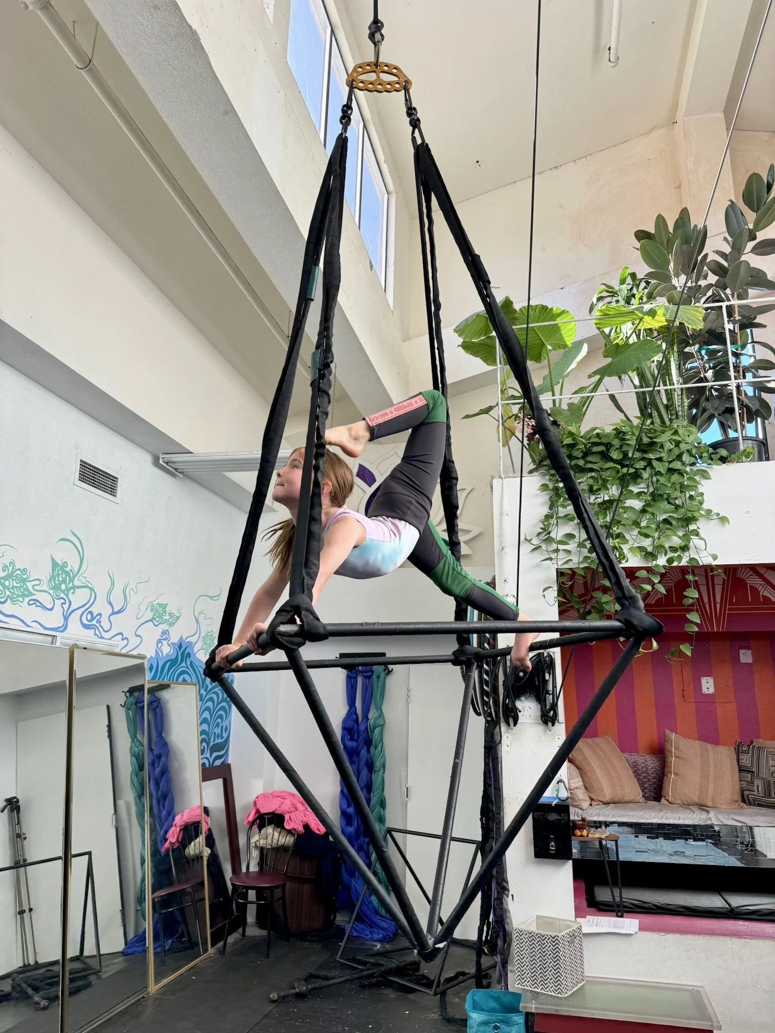 A young girl is performing an aerial yoga pose on a geometric aerial yoga hammock inside a room with plants, mirrors, and colorful decor.