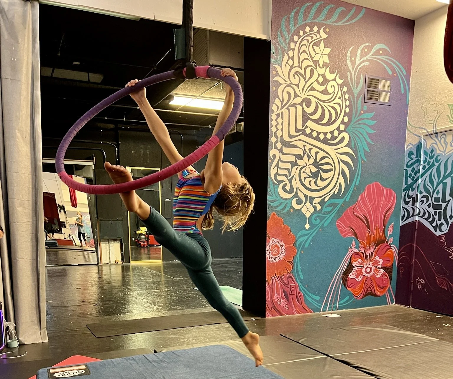 A woman practicing aerial acrobatics on a hoop in a colorful gym. She is holding the hoop with one hand, with her body extended horizontally and one leg bent upward. The gym features vibrant wall art with floral and swirling patterns.