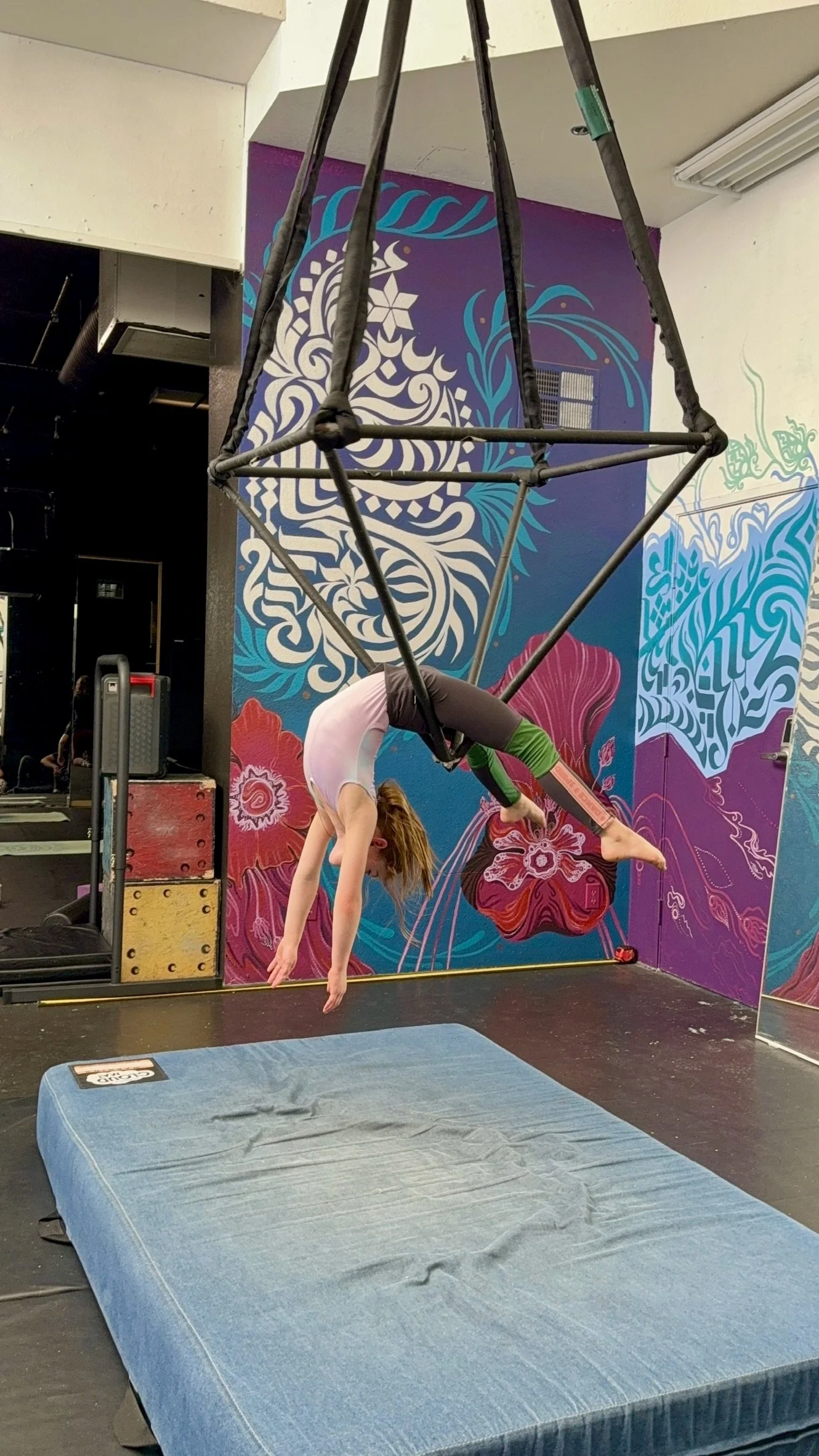A woman practicing aerial silk or hammock acrobatics in a gym with colorful mural art on the walls, with a blue crash pad below her