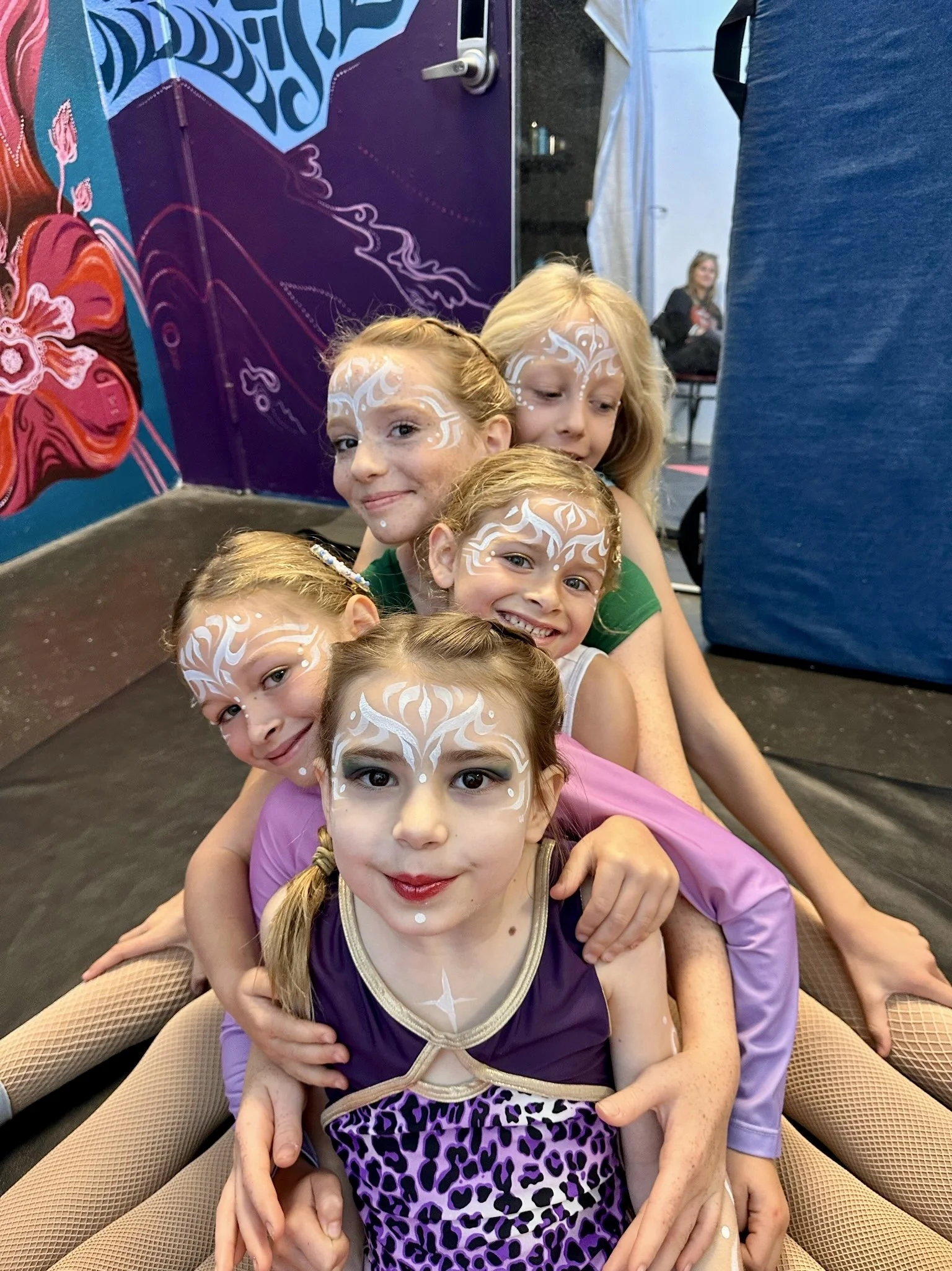 Five young girls with painted faces, sitting in a line, smiling and dressed in colorful costumes, at a performance or event with mural art and a blue padded wall in the background.
