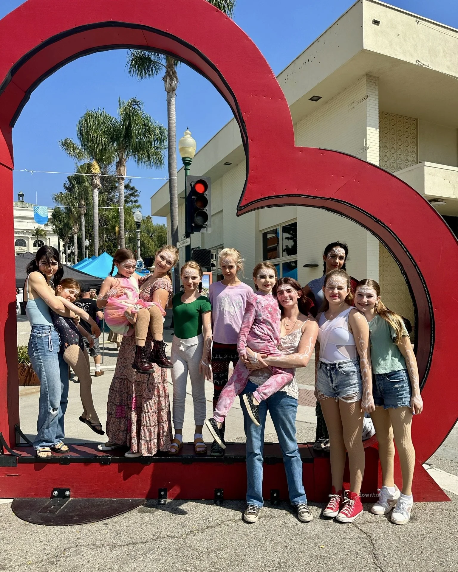 A group of children and adults posing inside and around a large red sculpture of a heart during an outdoor event on a sunny day, with palm trees and buildings in the background.