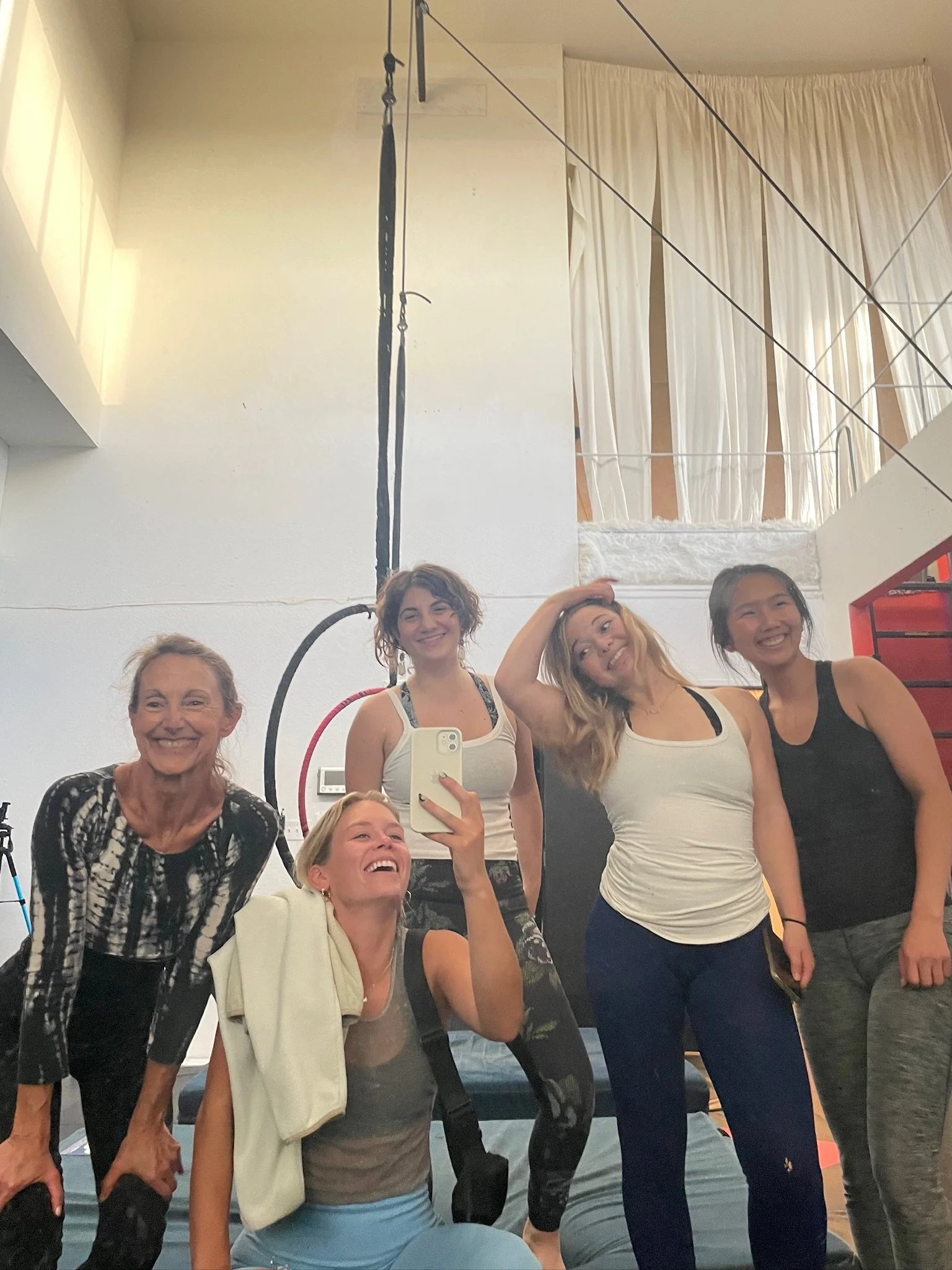 A group of five women taking a selfie in a fitness or gym studio, with gym equipment and climbing ropes in the background.