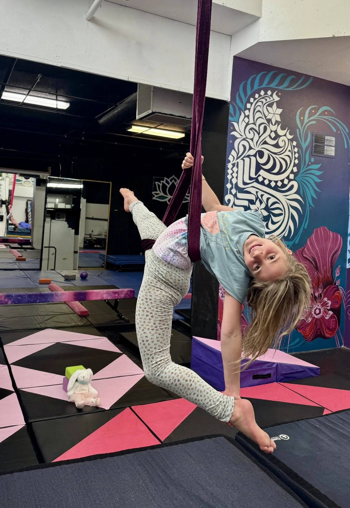 A young girl practicing aerial silk arts in a gym, hanging upside down and smiling, with colorful indoor gym equipment and a mural in the background.