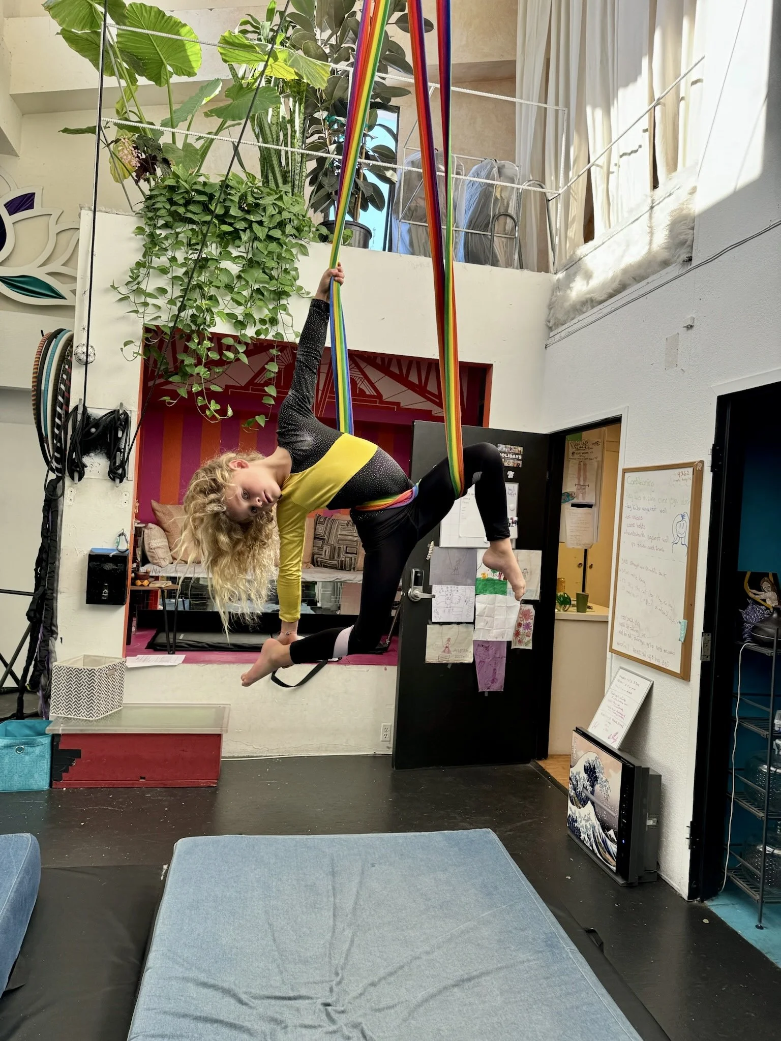 A young woman practicing aerial silks indoors, hanging upside down with her body bent and her head tilted to the side. She is holding onto rainbow-colored fabric with one hand, while her other arm supports her body. The room features plants, workout 