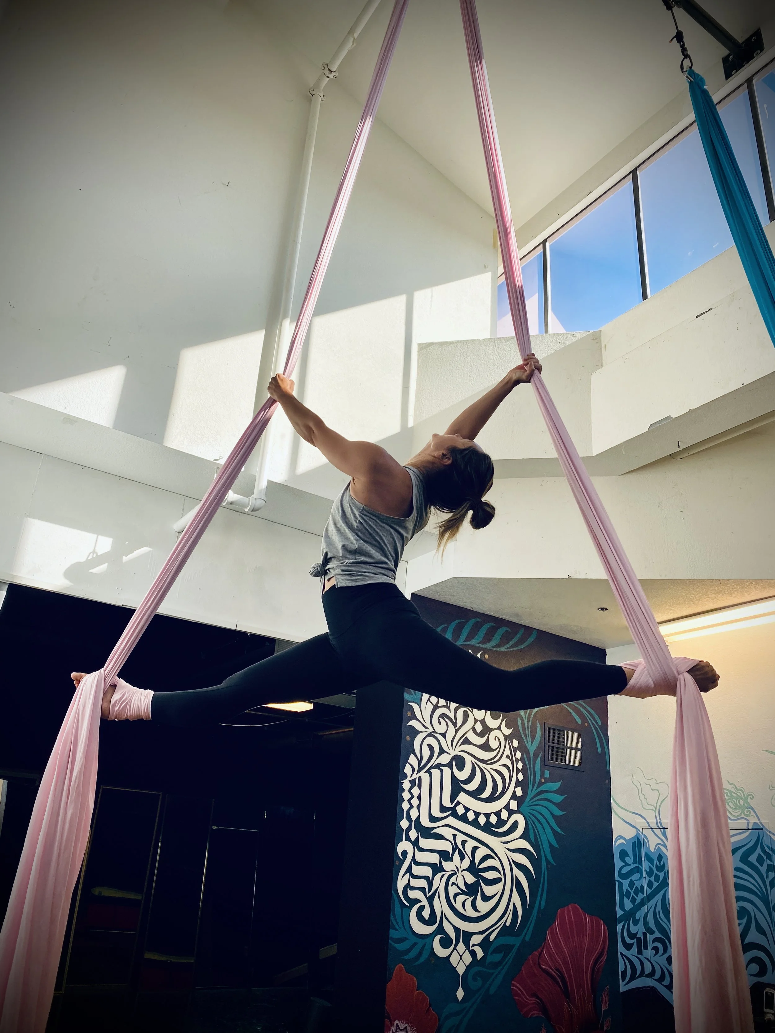 A woman practicing aerial silk acrobatics indoors, performing a split while hanging from pink fabric suspended from the ceiling.