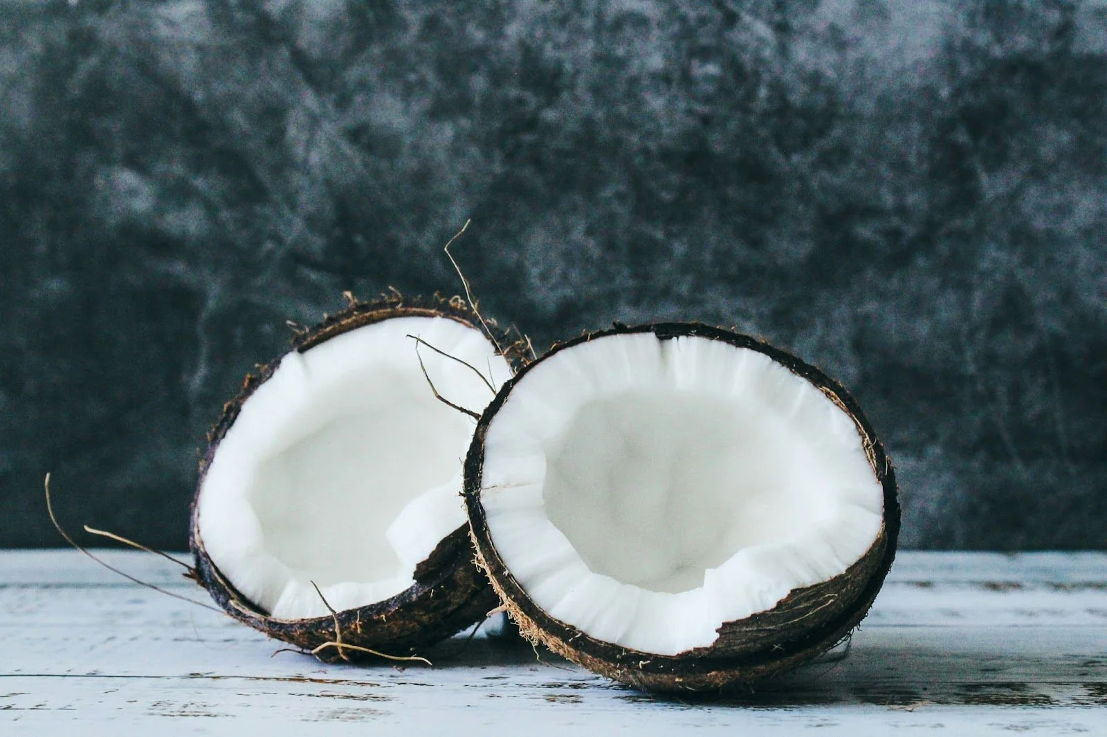 Two halves of a coconut with white flesh on a rustic white wooden surface, dark textured background.