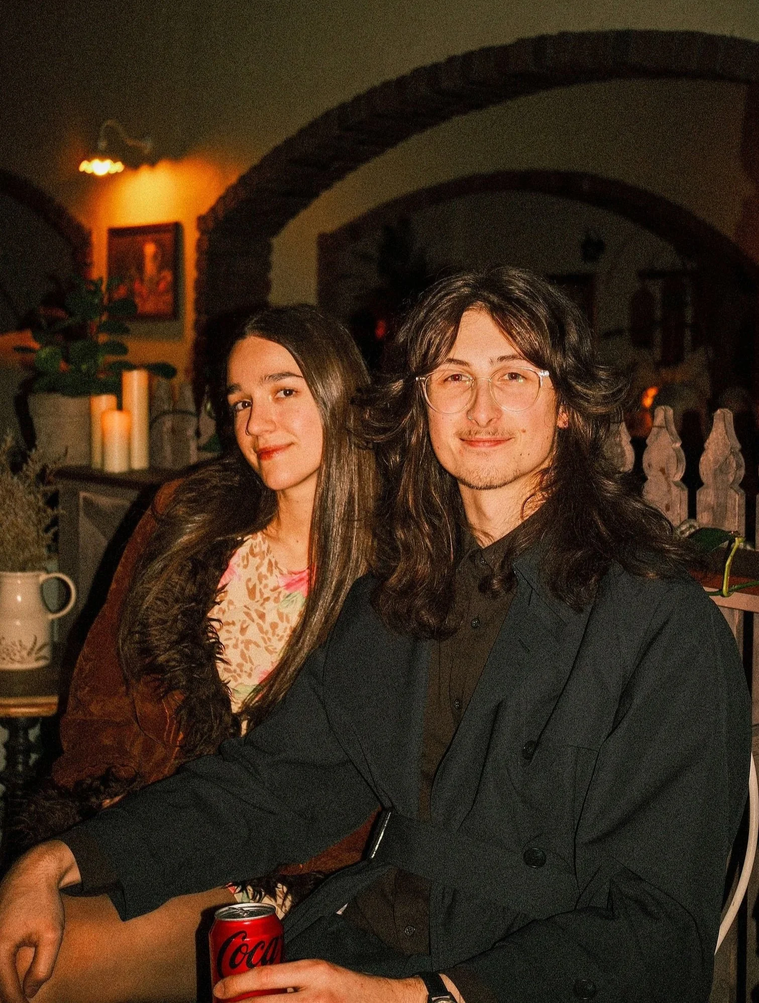 Owner of Loftes Photography, Tom Wilkinson and his partner, his partner is smiling and he is holding a Coca-Cola can, in a dimly lit room decorated with candles and potted plants.