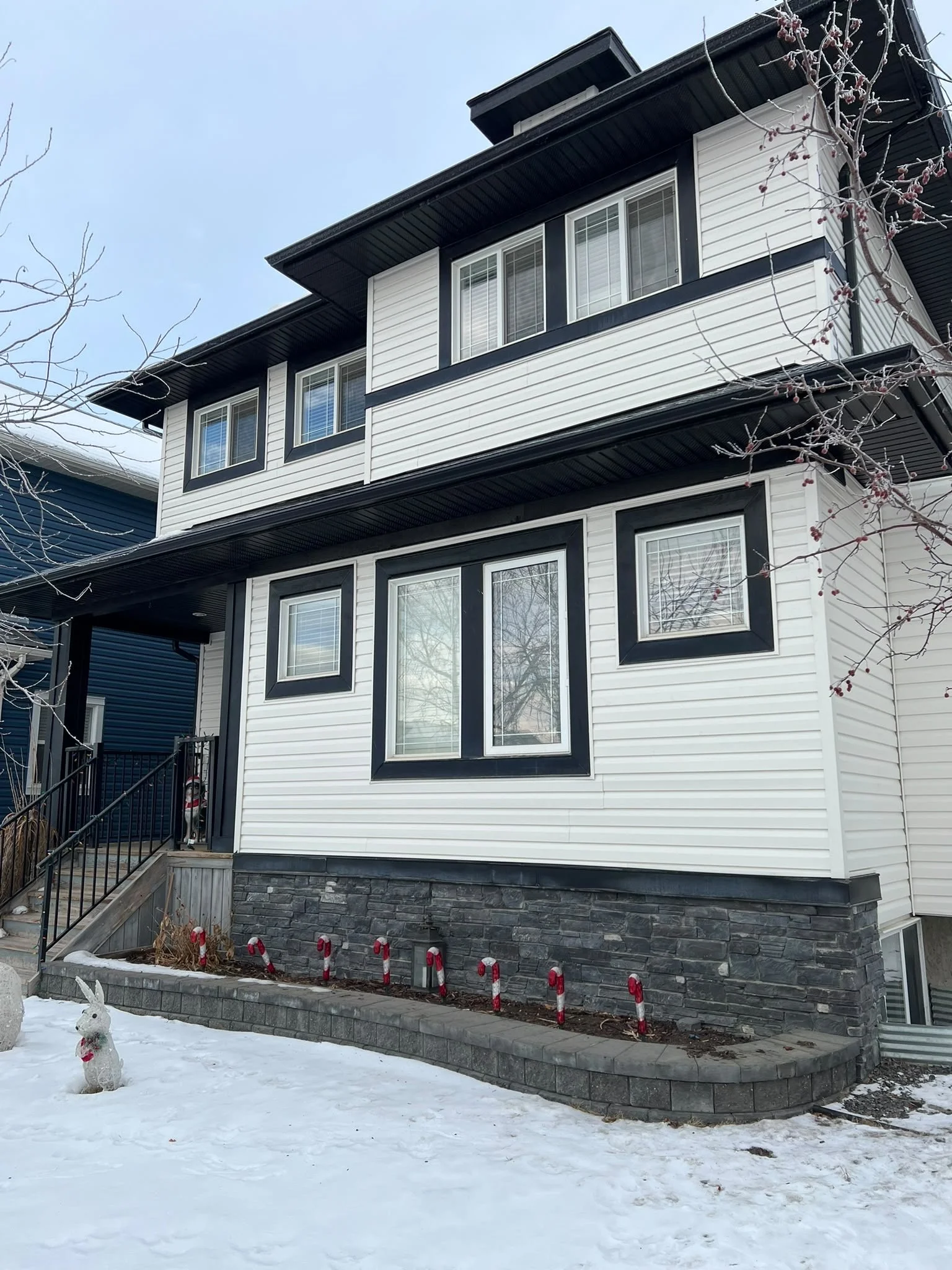 Front view of a modern, multi-story house with white and black exterior siding, multiple windows, and a stone foundation. Snow covers the ground, with a small decorative bunny figure in the snow, and a row of red and white candy cane garden stakes along a landscaped area.
