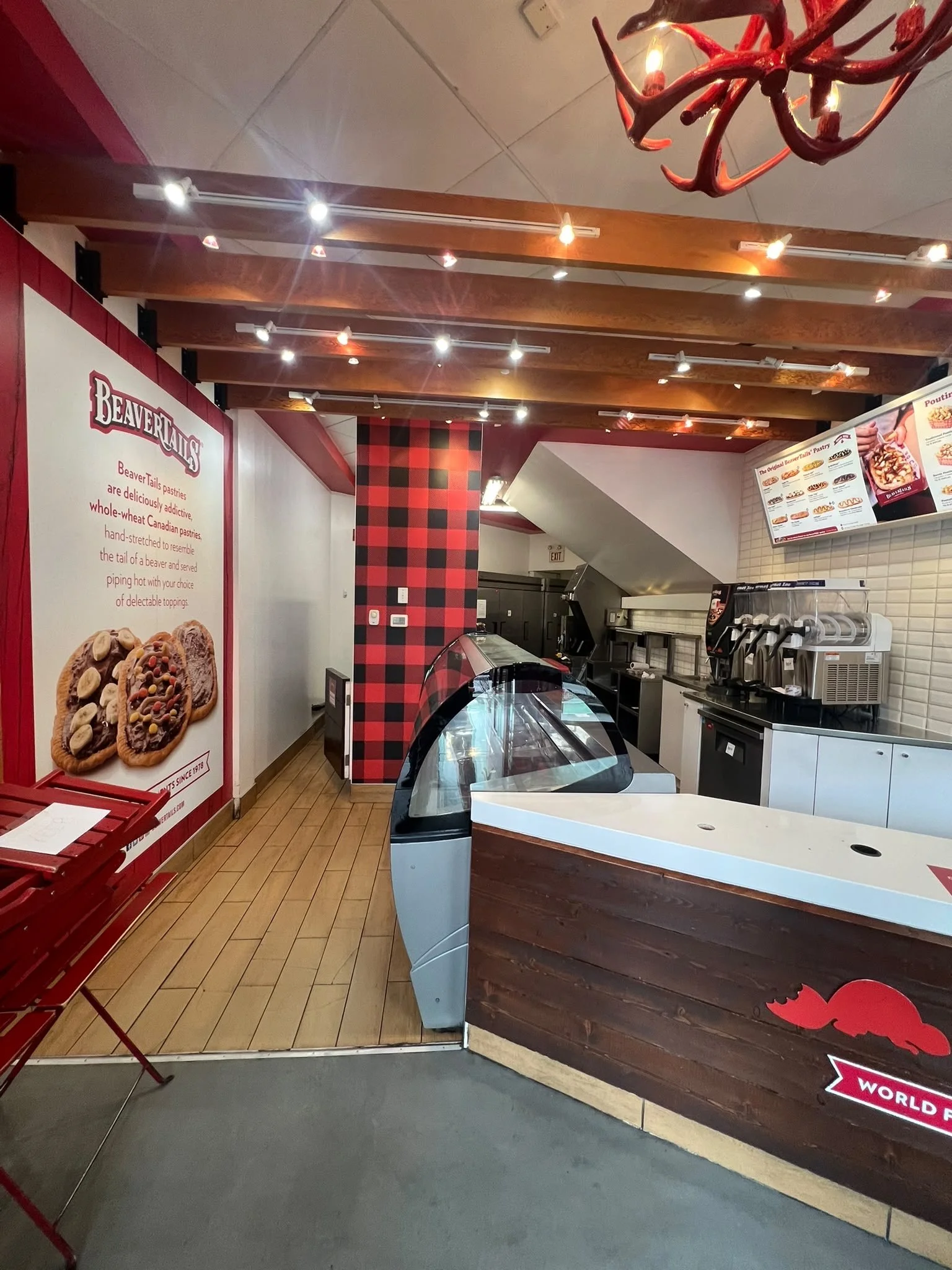Interior view of a bakery or pastry shop with a wooden and brick display counter, menu boards on the wall, and red plaid decor. There is a corner with a mosaic black and red checkered pattern near the ceiling.