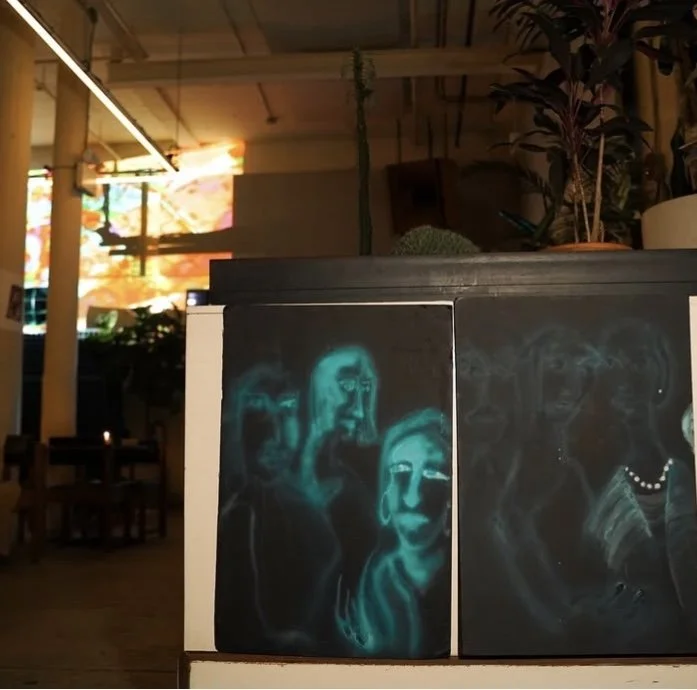 Glow-in-the-dark paintings of women with long hair on a dark background displayed on a white surface inside a dimly lit cafe or gallery. MARIAM KVASHILAVA EXHIBITION