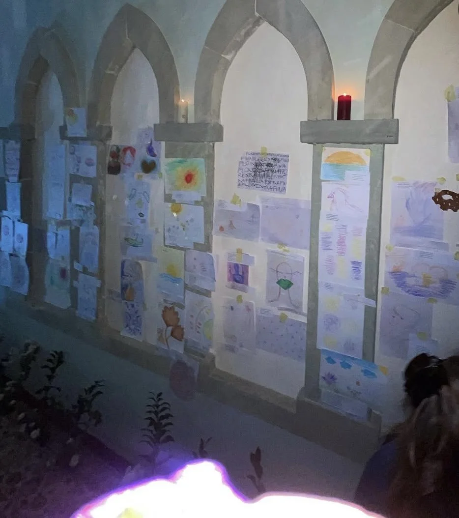 Wall decorated with children's drawings and writings, illuminated by candles on top of stone arches, with some plants visible at the bottom.