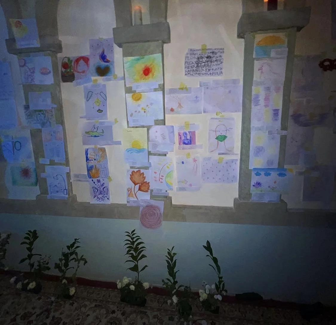 A wall decorated with children's colorful drawings and paintings, taped in a grid pattern. There are four concrete columns along the wall and potted plants with white flowers lined up at the bottom on a patterned rug.