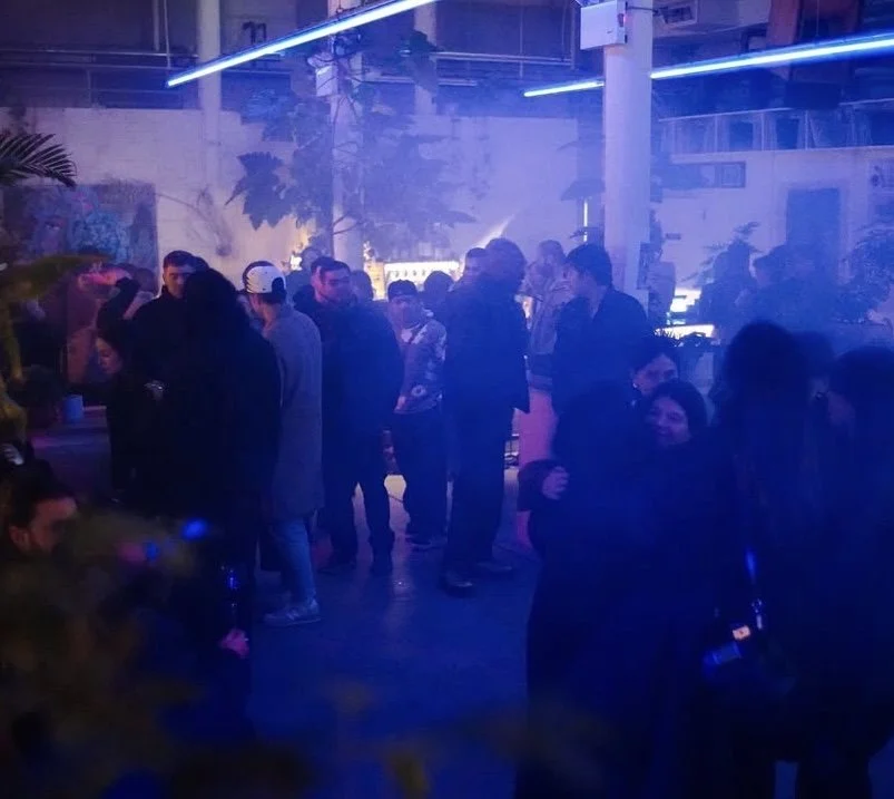 People gathered at a dimly lit indoor party or nightclub with blue neon lighting, some socializing and dancing. MARIAM KVASHILAVA EXHIBITION