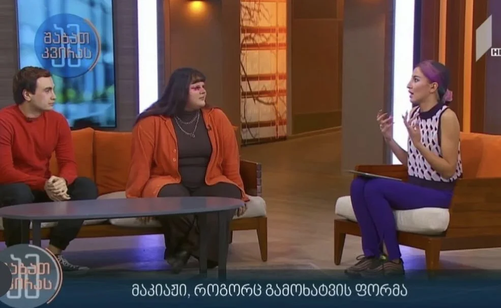 A teMARIAM KVASHILAVA talk show set with three women seated on chairs. The woman on the right is speaking, with purple hair and a patterned top, while two women on the left listen attentively. The background includes a screen and studio decor.