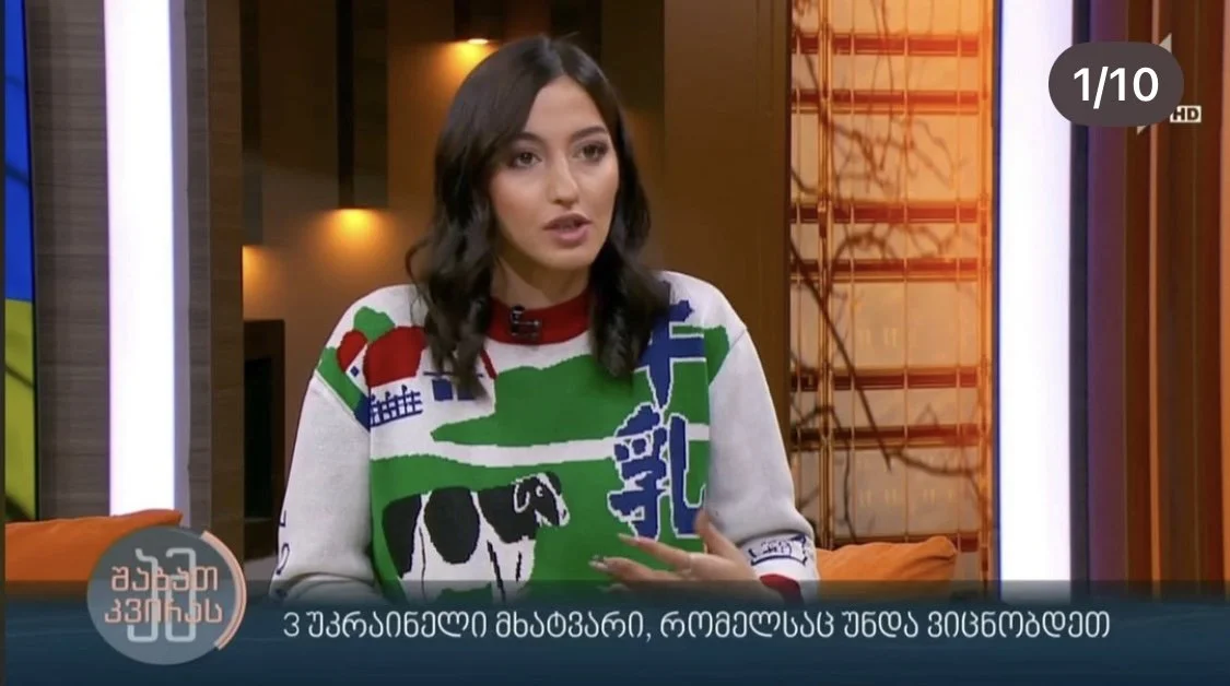 MARIAM KVASHILAVA with dark hair, wearing a colorful sweater, is speaking on a television set with Georgian text and graphics on the screen.