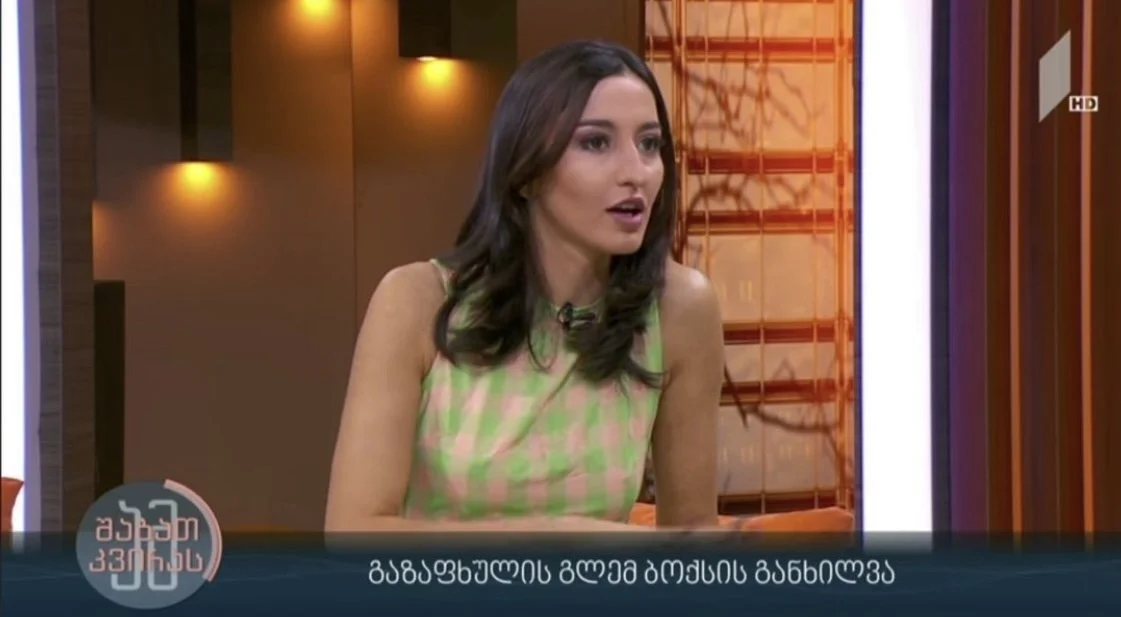 MARIAM KVASHILAVA Television talk show with a woman speaking, wearing a sleeveless, pink and green checkered dress, sitting in studio with warm lighting and wooden decor.
