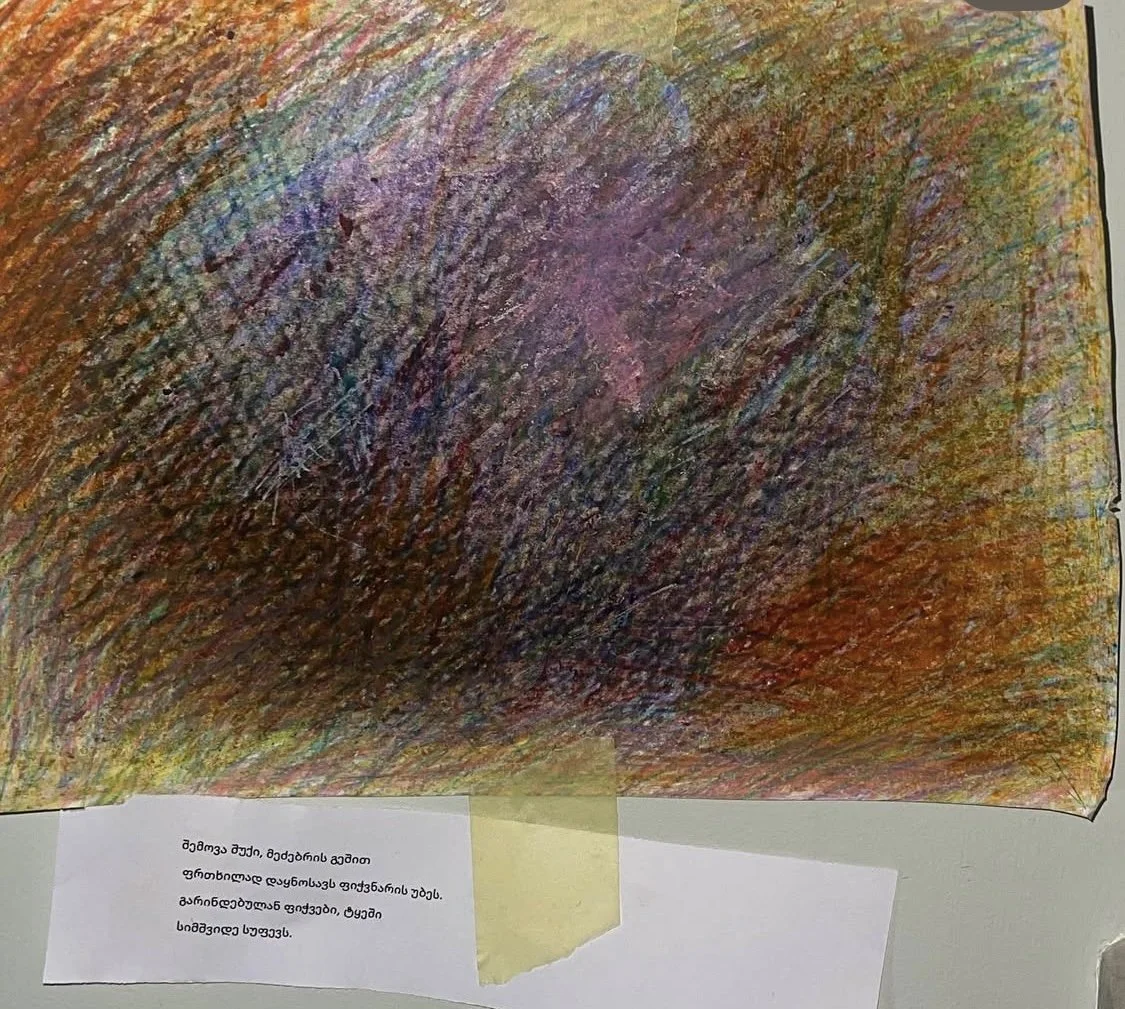 Child's crayon drawing on paper with scribbles in various colors, taped to a white surface alongside a piece of paper with text in a different language.